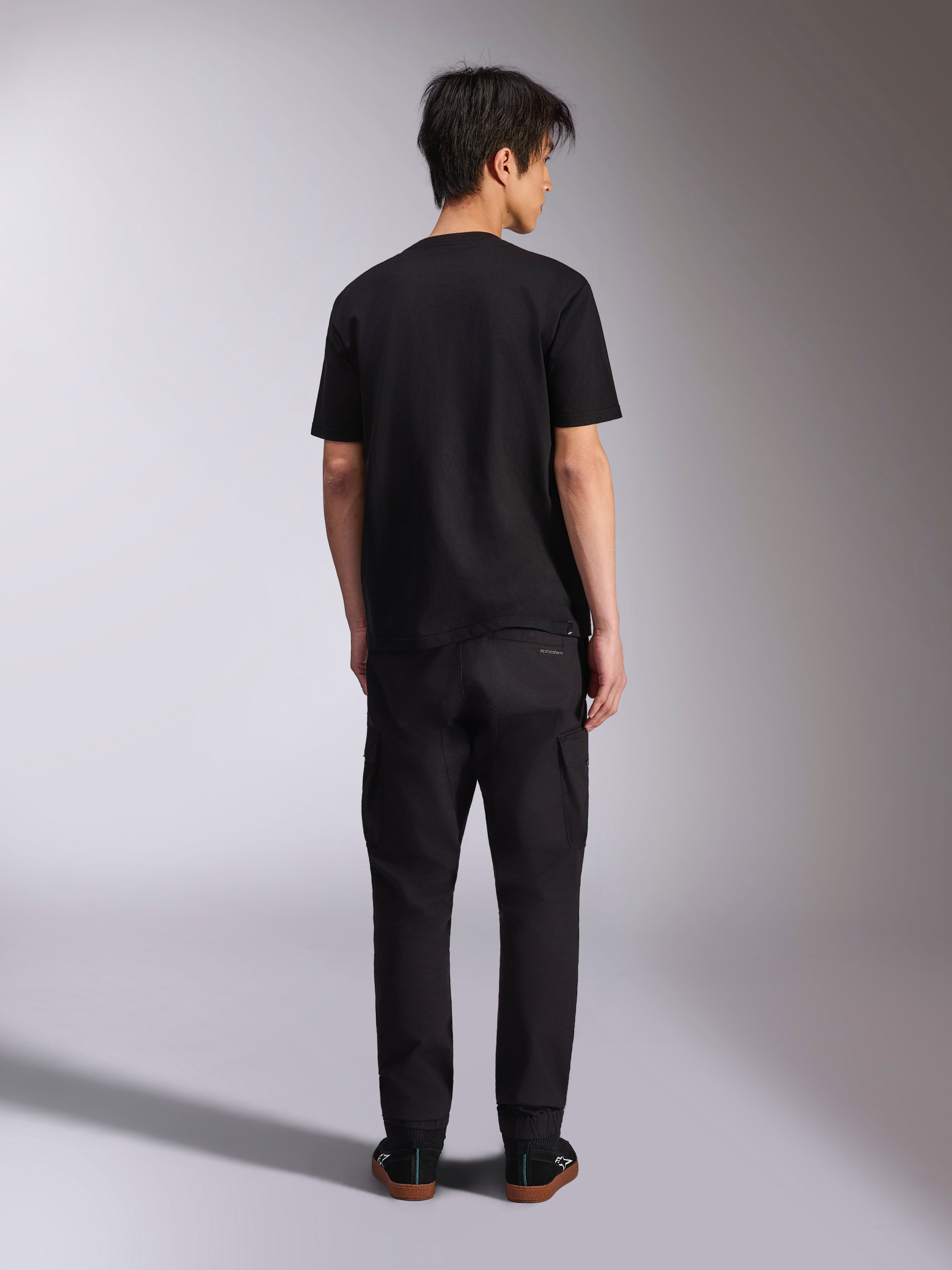 Alpinestars Noka CSF Tee, T-shirt met korte mouwen, zwart, achteraanzicht van model dat het shirt draagt met een zwarte broek en sneakers, casual motorsport streetwear-stijl