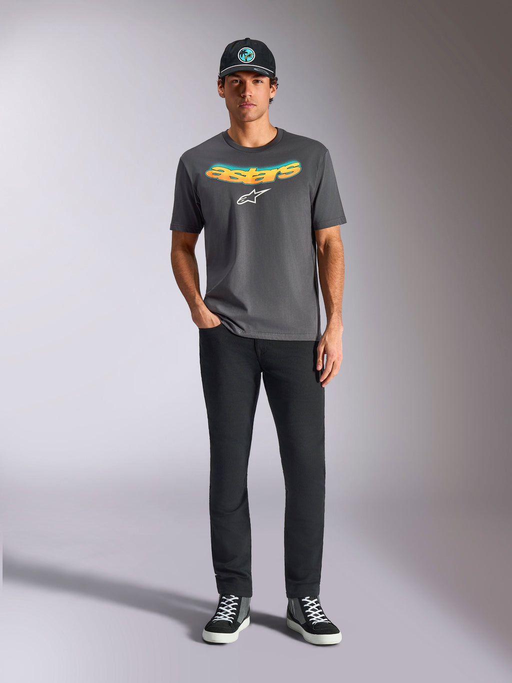 Alpinestars Ellipsoid CSF Tee, T-shirt met korte mouwen, antracietgrijs, met een gebogen geel en oranje kleurverloop tekstlogo en wit Astars-logo, gedragen door een model met zwarte broek en een baseballpet, casual sportkledingstijl