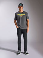 Alpinestars Ellipsoid CSF Tee, T-shirt met korte mouwen, antracietgrijs, met een gebogen geel en oranje kleurverloop tekstlogo en wit Astars-logo, gedragen door een model met zwarte broek en een baseballpet, casual sportkledingstijl