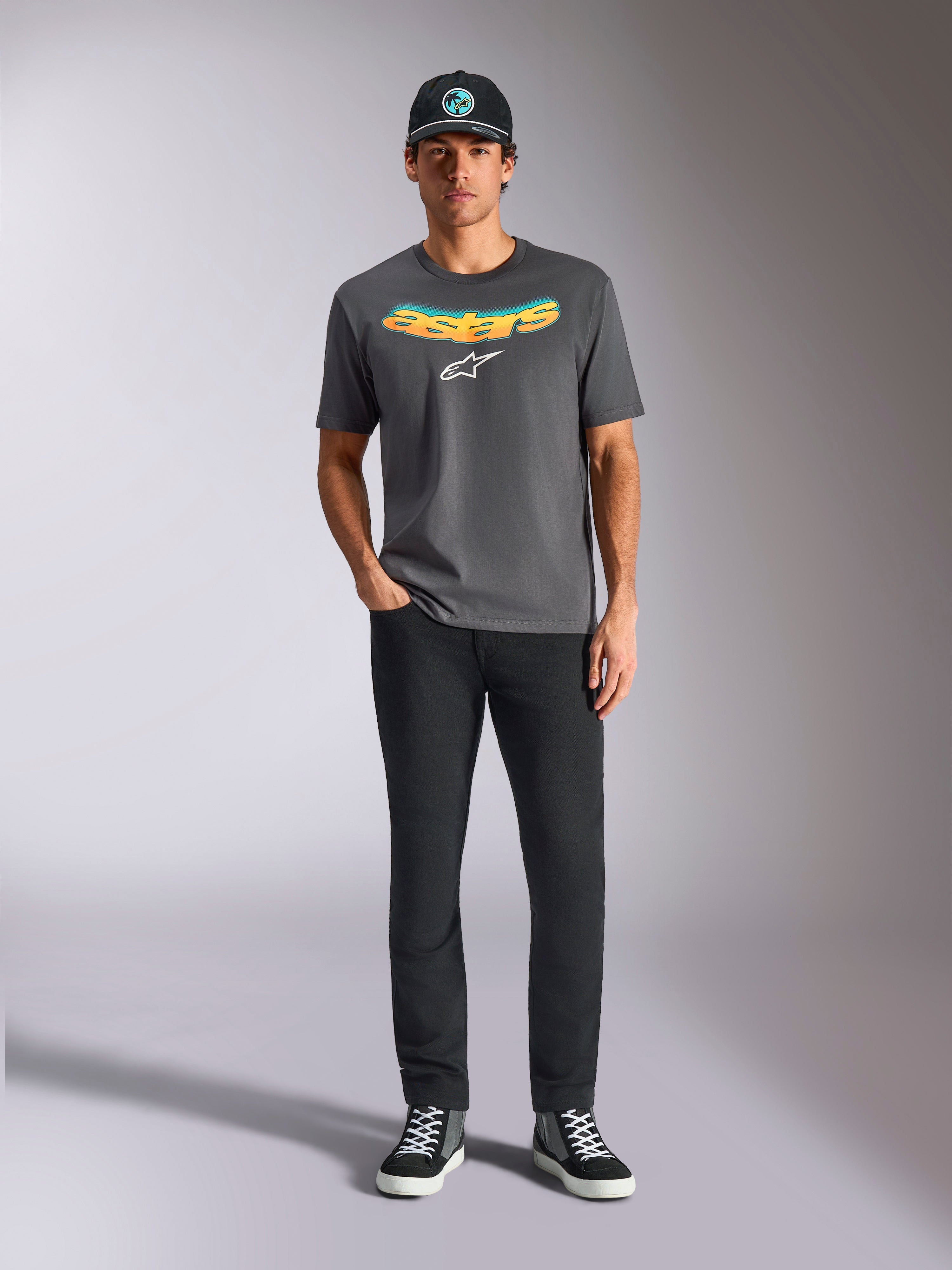 Alpinestars Ellipsoid CSF Tee, T-shirt met korte mouwen, antracietgrijs, met een gebogen geel en oranje kleurverloop tekstlogo en wit Astars-logo, gedragen door een model met zwarte broek en een baseballpet, casual sportkledingstijl