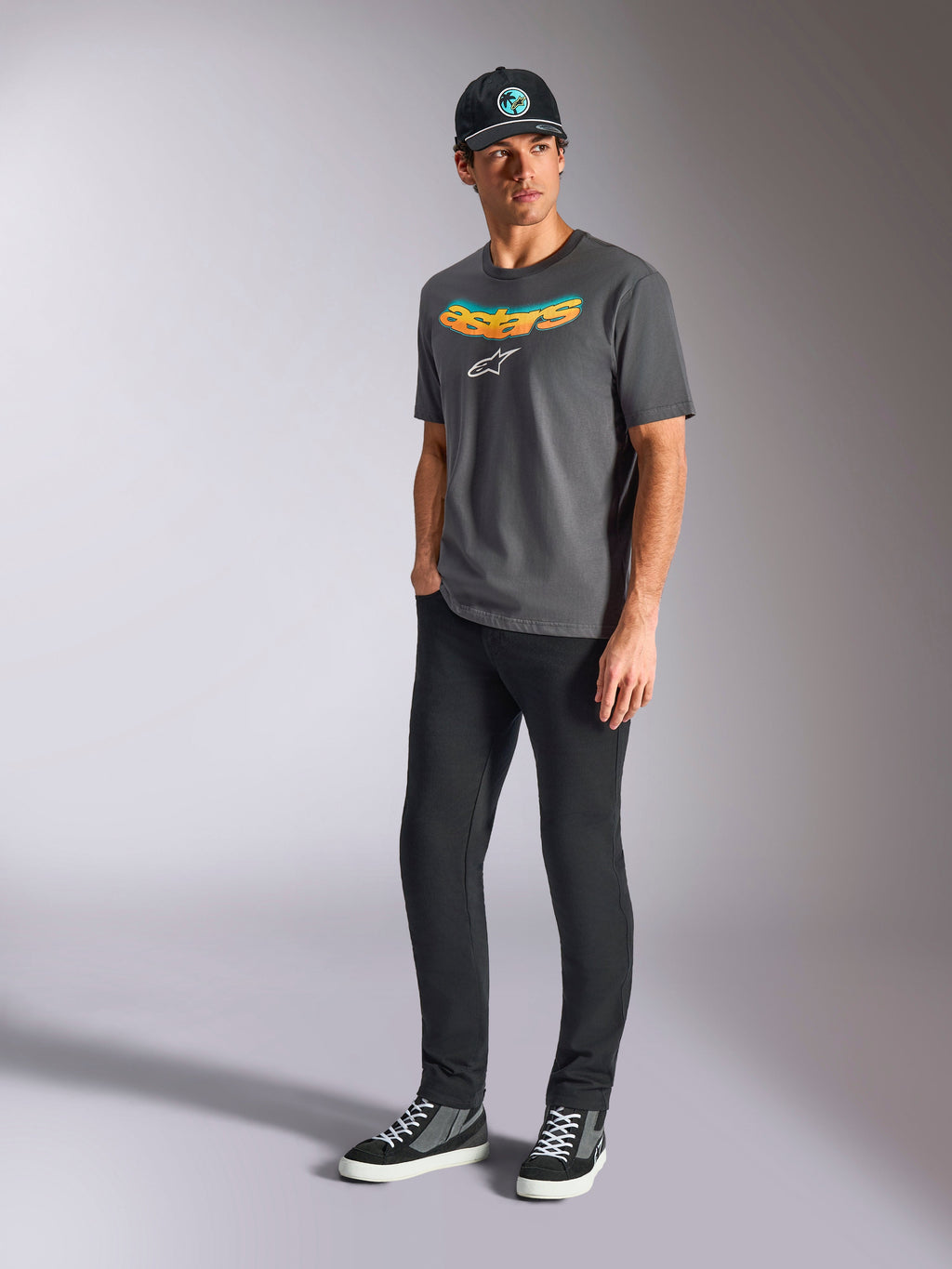 Alpinestars Ellipsoid CSF Tee, T-shirt met korte mouwen, antracietgrijs, met gebogen Astars-woordmerk met kleurverloop en wit logo, gedragen door model met zwarte broek en pet, casual motorsport streetwear