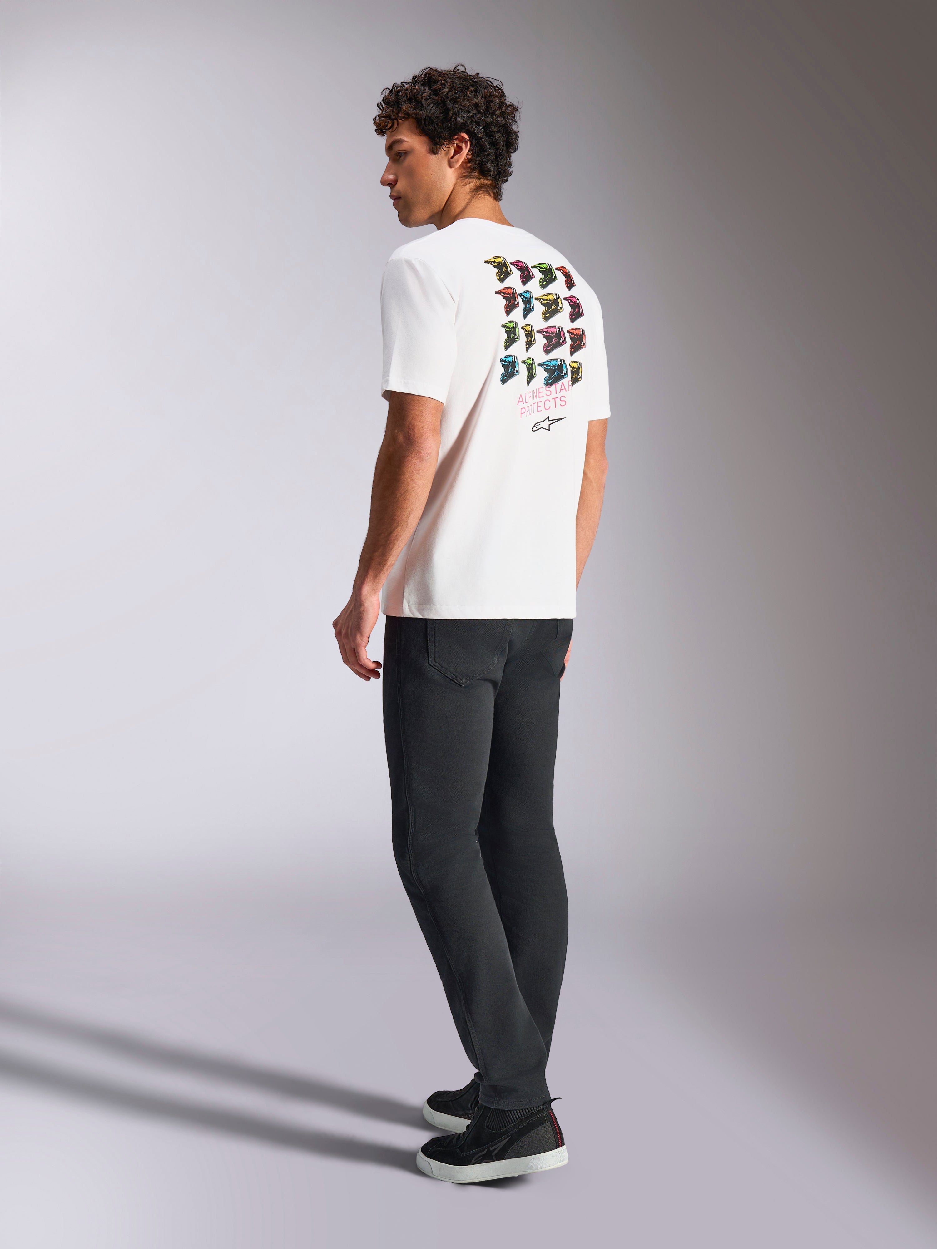 Alpinestars Pop Helmet CSF Tee, wit T-shirt met korte mouwen, achteraanzicht op model met kleurrijke M10 helm graphics, gestyled met een donkere broek en zwarte sneakers, casual motorsport-geïnspireerde streetwear