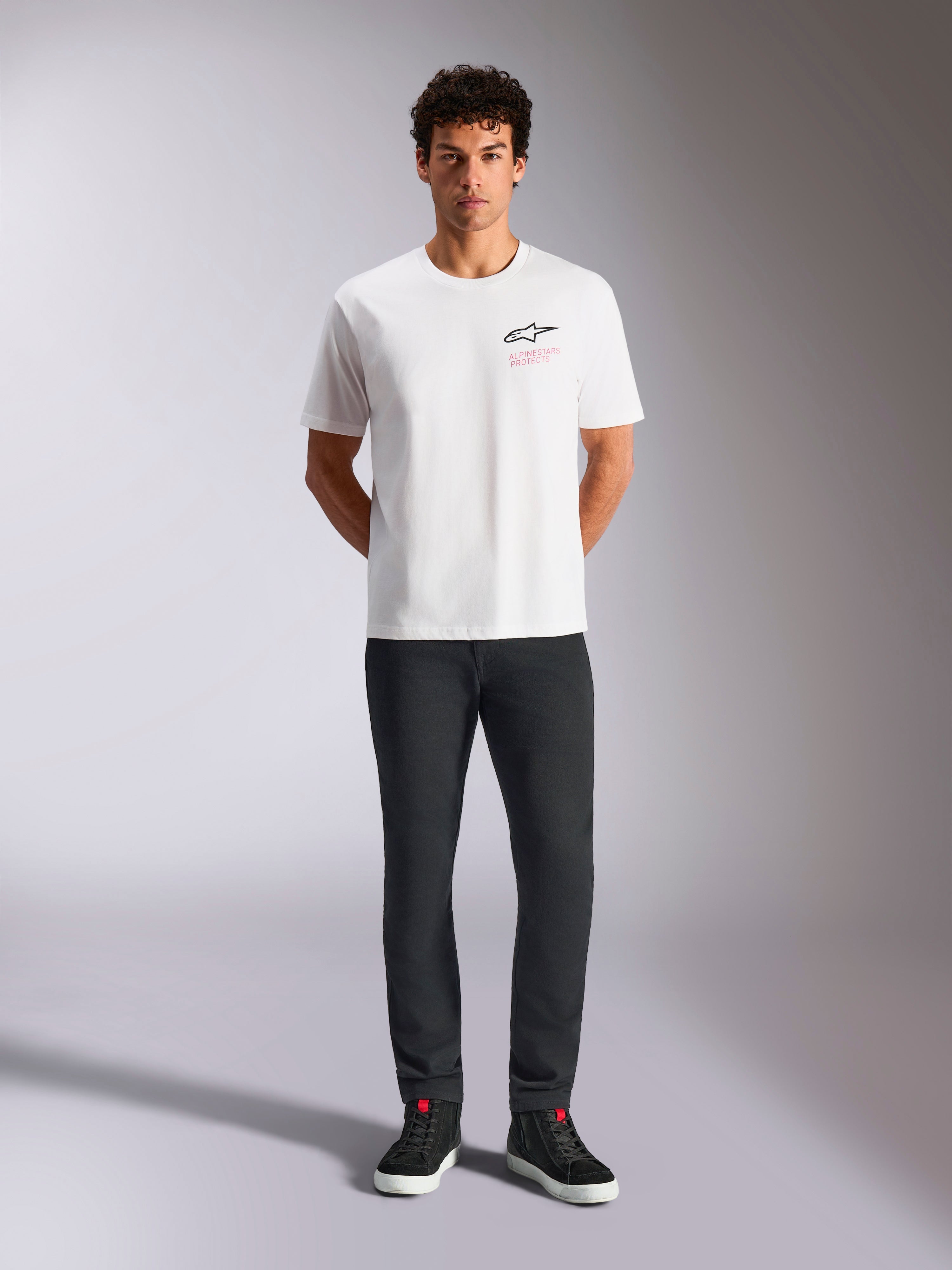 Alpinestars Pop Helmet CSF Tee, T-shirt met korte mouwen, wit, gedragen door een model met een zwarte broek en zwarte high-top sneakers, Astars-logo op de voorzijde met roze 'Alpinestars Protects' tekst, casual motorsport streetwear