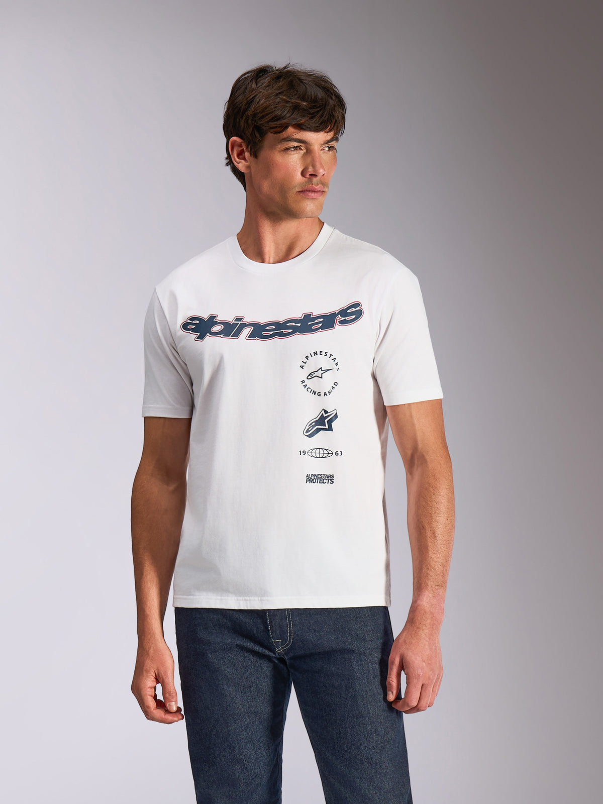 Alpinestars Abus CSF Tee, T-shirt met korte mouwen, wit, gebogen Alpinestars-logo met meervoudige gestapelde graphics op de borst, gedragen door model met donkere denim jeans, casual motorsport streetwear