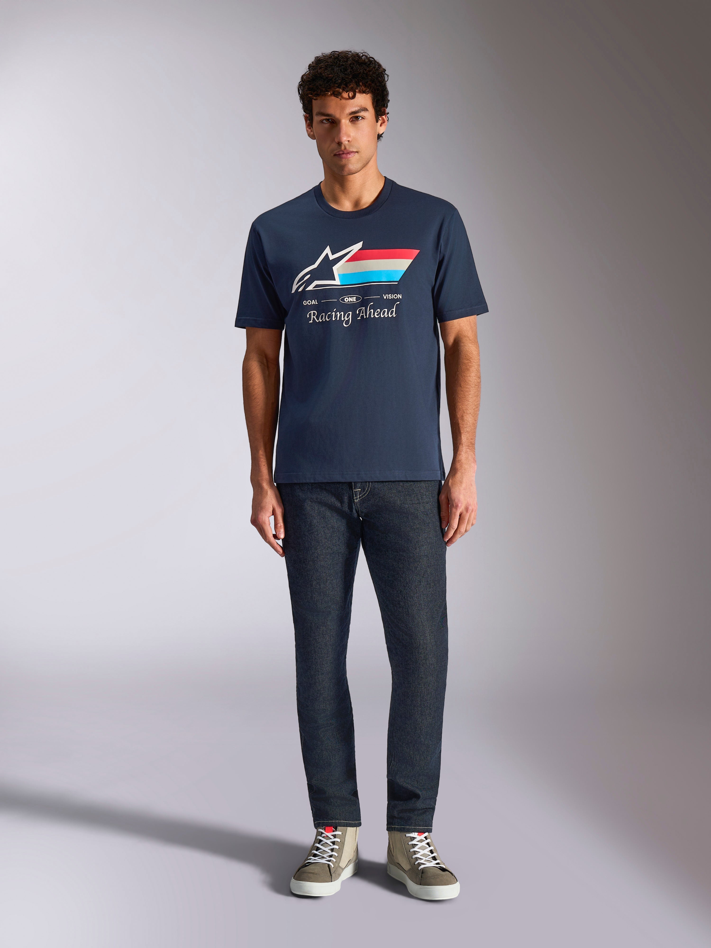 Alpinestars Trifecta CSF T-shirt met korte mouwen, marineblauw, wit Astars-logo met rode, witte en lichtblauwe strepen en 'Racing Ahead' tekst, gedragen met donkere jeans en olijfgroene sneakers, casual motorsport streetwear
