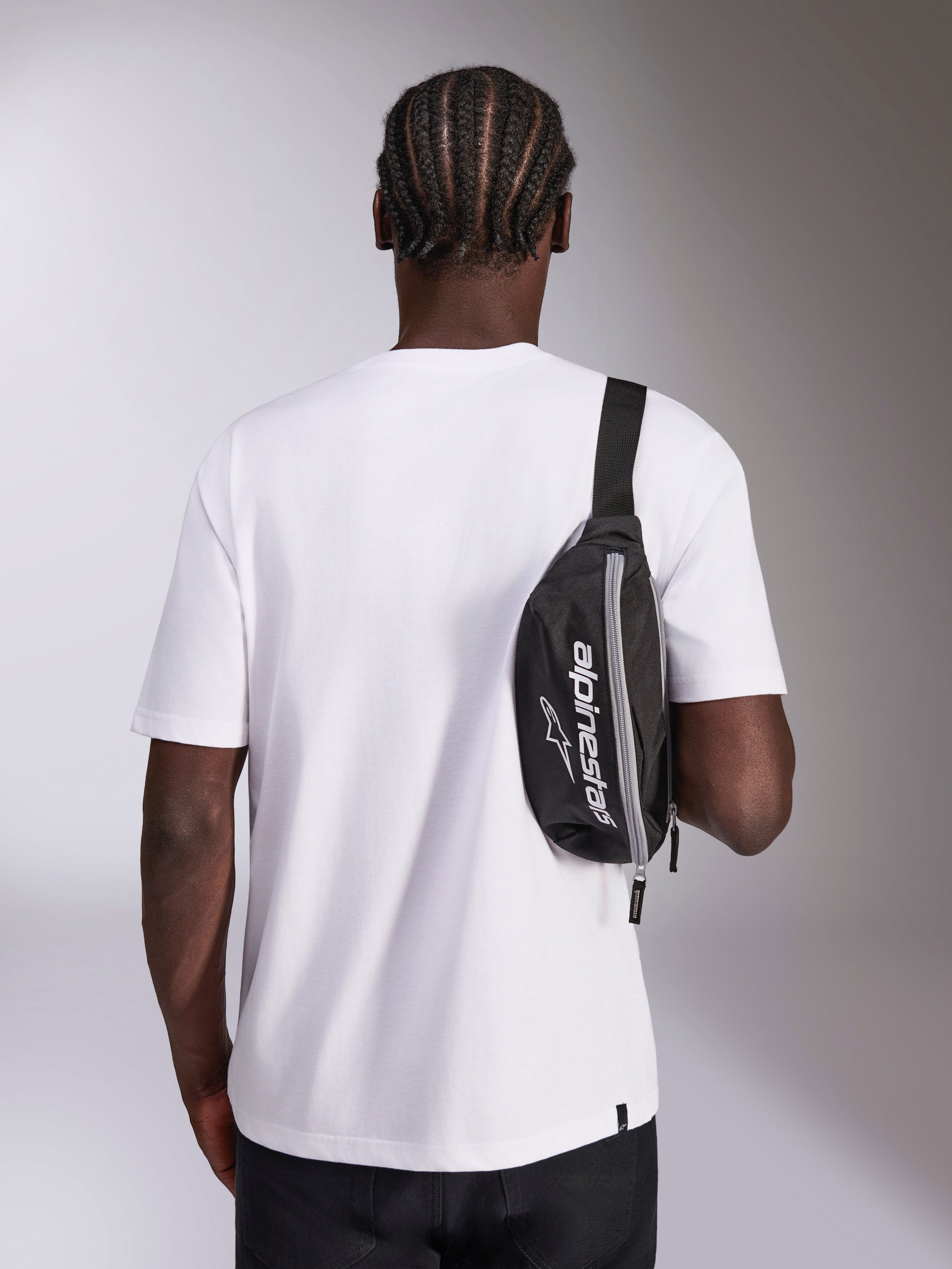 Alpinestars Only Linear heuptas, zwart met wit Alpinestars-logo, gedragen als crossbodytas over een wit T-shirt, casual motorsport-geïnspireerd streetwear-accessoire