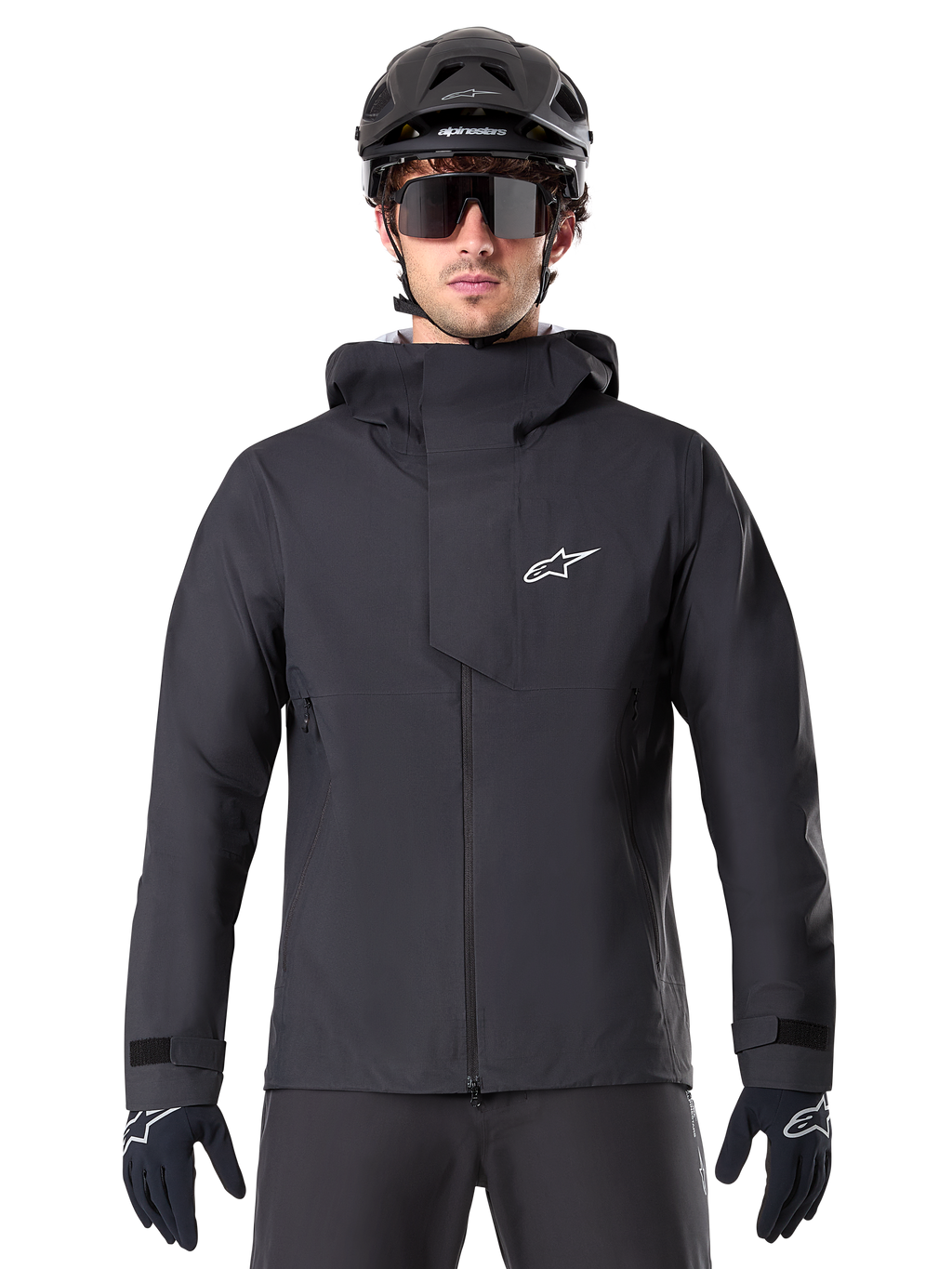 Alpinestars A-Dura Elite Waterproof Jacket, zwarte mountainbike jas, hoge kraag met capuchon, ritssluiting aan de voorzijde met beschermende borstflap en wit logo, gedragen met zwarte helm en fietshandschoenen, technische weersbescherming voor trailrijden