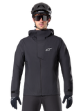 Alpinestars A-Dura Elite Waterproof Jacket, zwarte mountainbike jas, hoge kraag met capuchon, ritssluiting aan de voorzijde met beschermende borstflap en wit logo, gedragen met zwarte helm en fietshandschoenen, technische weersbescherming voor trailrijden