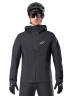 Alpinestars A-Dura Elite Waterproof Jacket, zwarte mountainbike jas, hoge kraag met capuchon, ritssluiting aan de voorzijde met beschermende borstflap en wit logo, gedragen met zwarte helm en fietshandschoenen, technische weersbescherming voor trailrijden