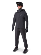 Alpinestars A-Dura Elite waterdichte jas in het zwart, gedragen door een mannelijke rider met bijpassende zwarte MTB-broek, handschoenen en helm. De jas heeft een technisch waterdicht ontwerp met een wit Astars-logo op de borst en een hoge beschermende kraag.