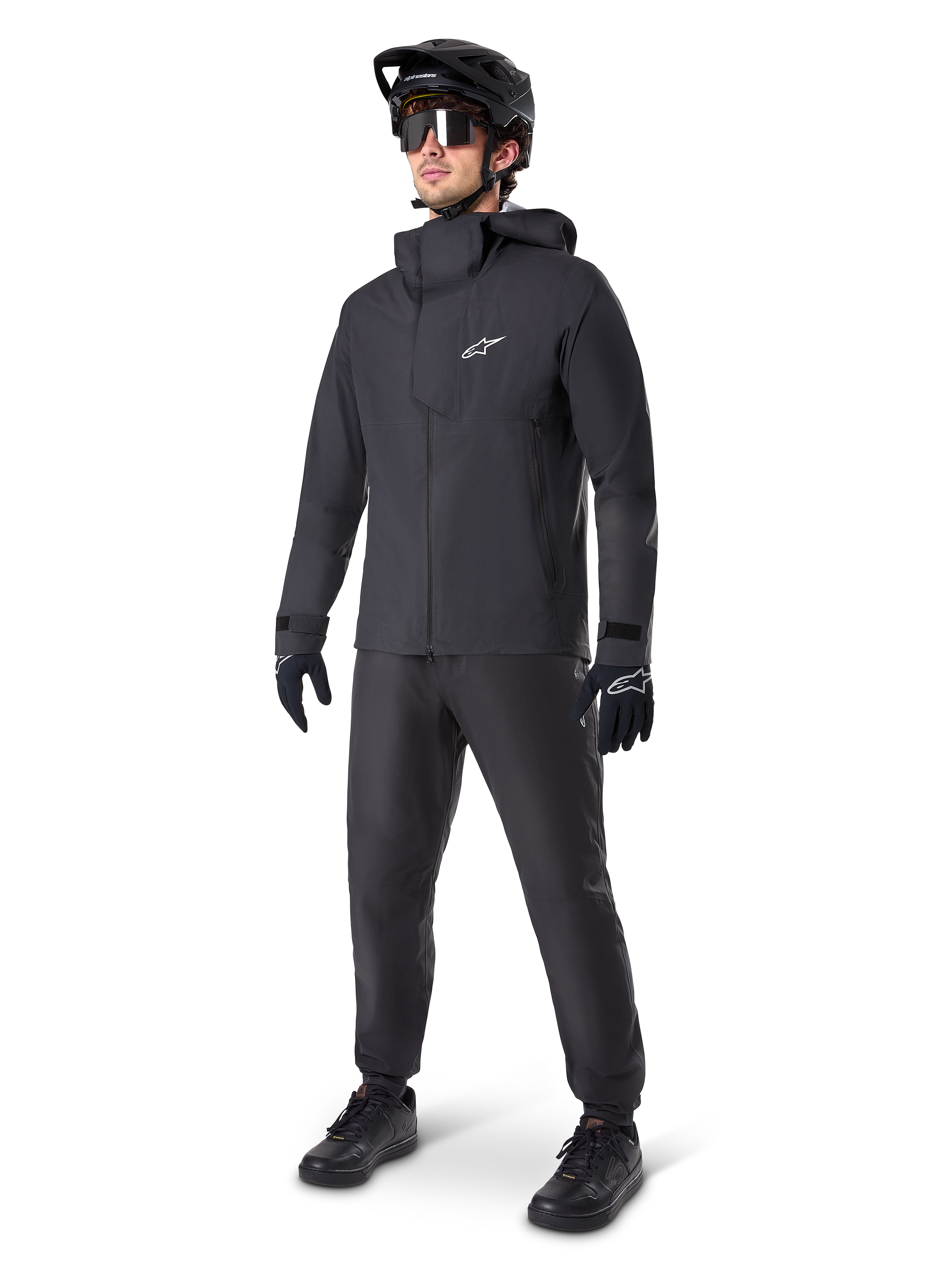 Alpinestars A-Dura Elite waterdichte jas in het zwart, gedragen door een mannelijke rider met bijpassende zwarte MTB-broek, handschoenen en helm. De jas heeft een technisch waterdicht ontwerp met een wit Astars-logo op de borst en een hoge beschermende kraag.