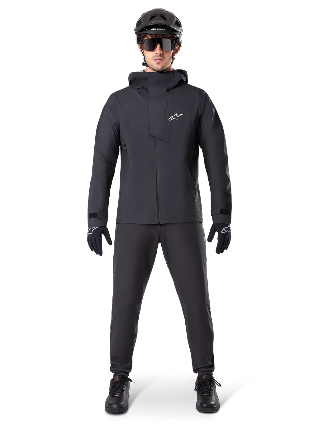 Alpinestars A-Dura Elite waterdichte jas, zwart, gedragen met bijpassende fietsbroek en handschoenen, zwarte helm en zonnebril, voorzien van een wit Astars-logo op de borst, hoogwaardig technisch ontwerp voor mountainbiken