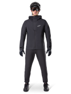 Alpinestars A-Dura Elite waterdichte jas, zwart, gedragen met bijpassende fietsbroek en handschoenen, zwarte helm en zonnebril, voorzien van een wit Astars-logo op de borst, hoogwaardig technisch ontwerp voor mountainbiken