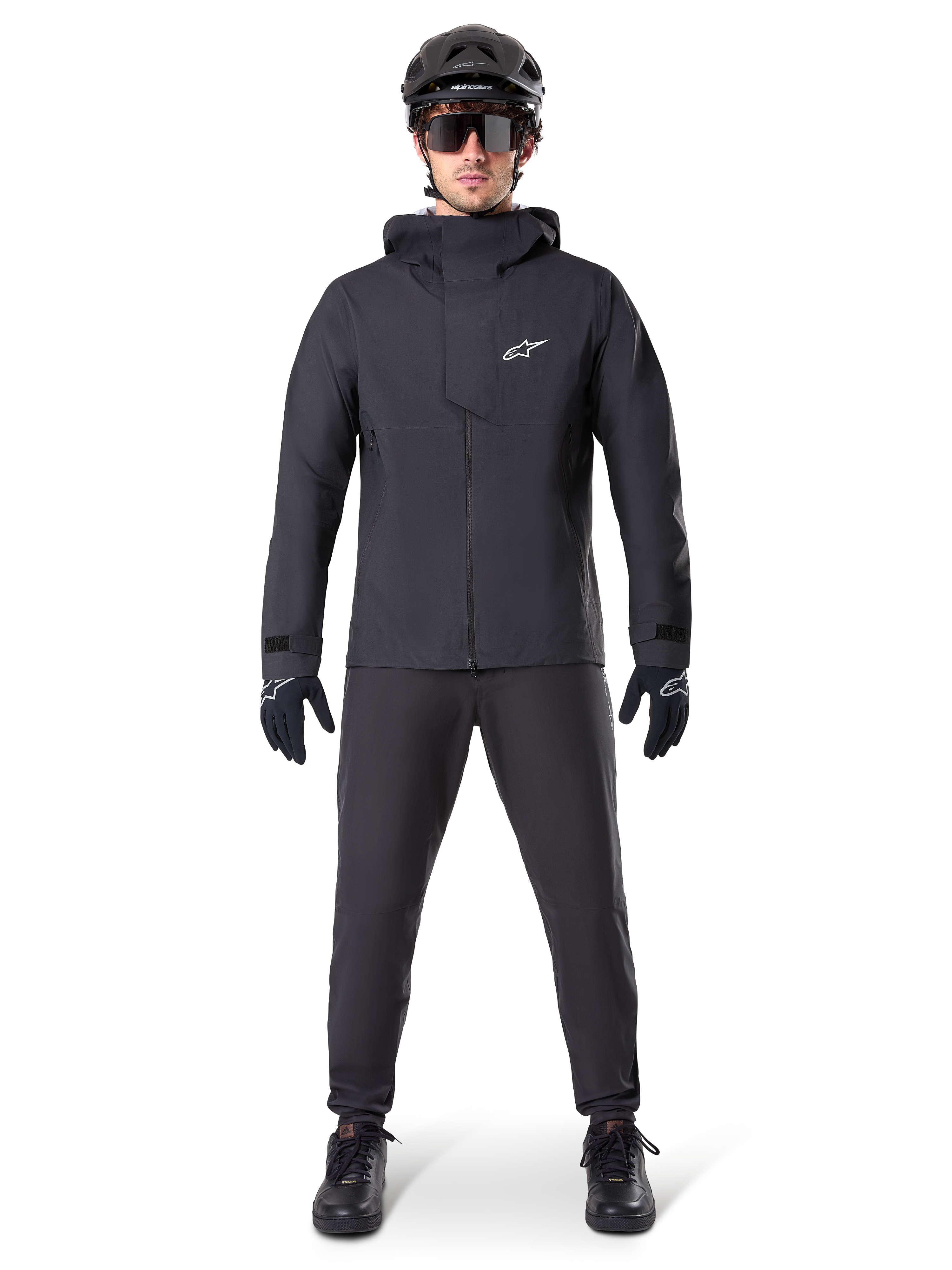 Alpinestars A-Dura Elite waterdichte jas, zwart, gedragen met bijpassende fietsbroek en handschoenen, zwarte helm en zonnebril, voorzien van een wit Astars-logo op de borst, hoogwaardig technisch ontwerp voor mountainbiken