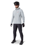 Alpinestars A-Dura Thermal Jacket in Titanium, light gray hooded cycling jacket with a red Alpinestars logo on the chest, worn by a man with a black MTB helmet, sunglasses, dark gray pants, and black gloves, professional mountain bike gear