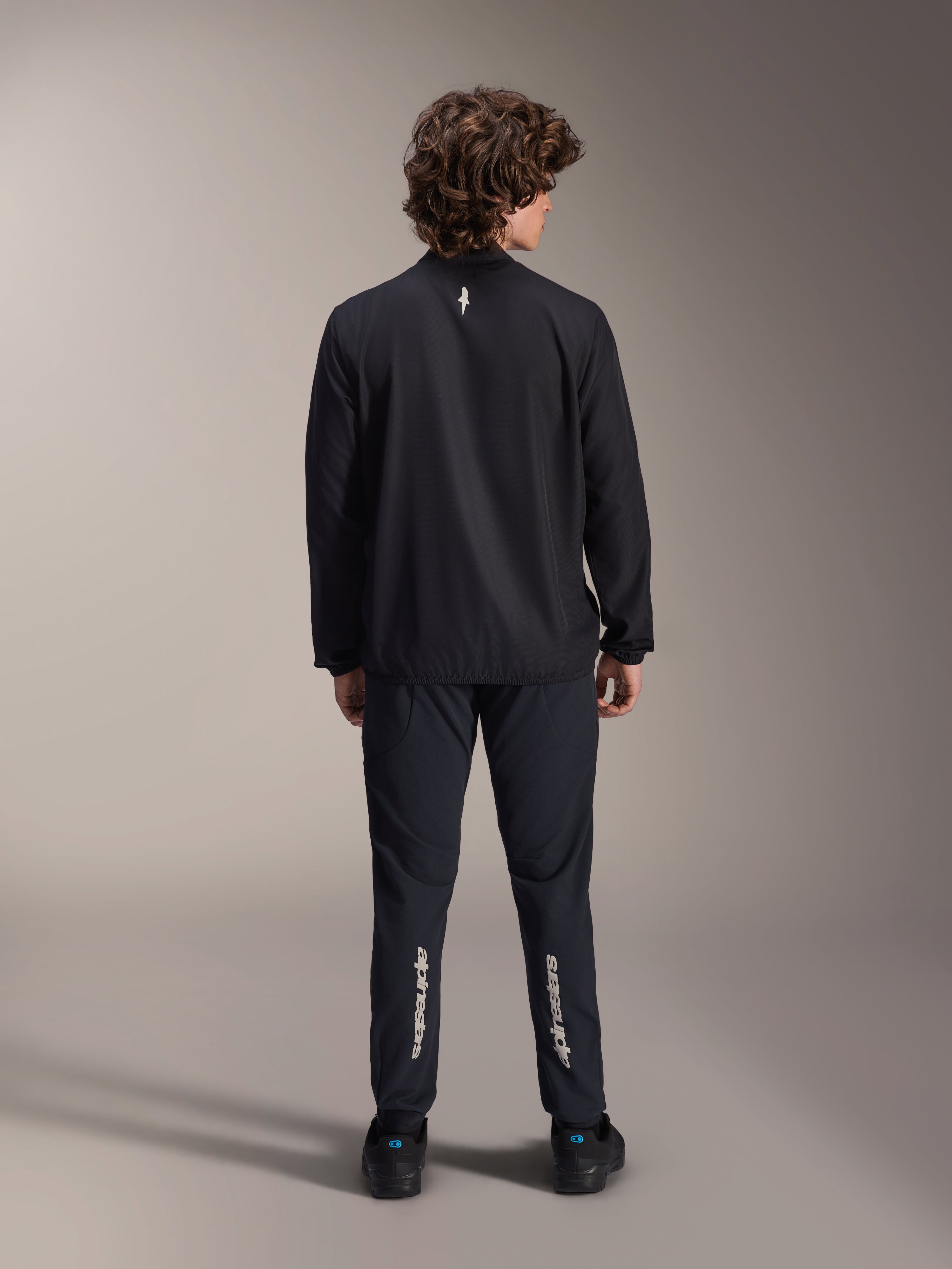 Alpinestars A-Aria Windbreaker Jacket, Black, back view, lightweight wind-resistant cycling jacket with a small white logo on the upper back, worn with matching black pants featuring vertical Alpinestars branding on the calves, performance mountain bike gear
