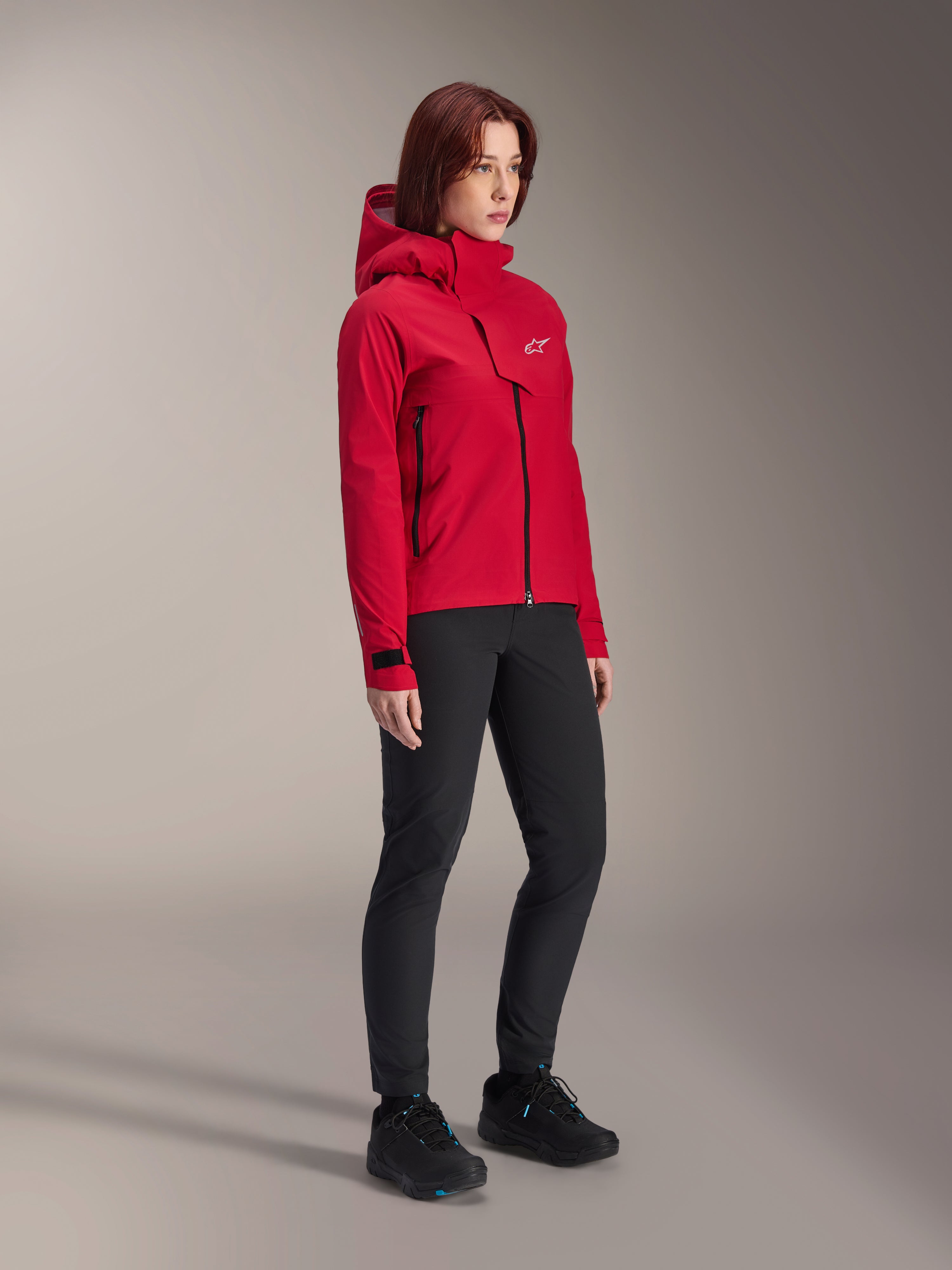 Alpinestars Stella A-Dura Elite waterdichte damesjas, Rood Fluo, mountainbike-regenjas met capuchon en wit Astars-logo op de borstflap, gedragen met zwarte broek en zwarte mountainbikeschoenen, technische prestatiekleding voor trailrijden