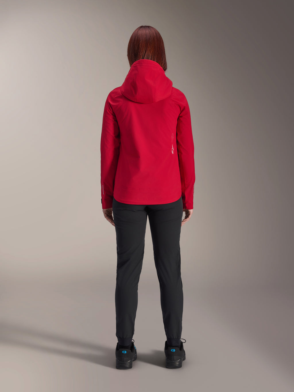 Achteraanzicht van een vrouw die de Alpinestars Stella A-Dura Elite Waterproof Jacket in Red Fluo draagt, gecombineerd met een zwarte broek en schoenen, voorzien van een beschermende capuchon en een verlengde achterzoom voor mountainbiken.