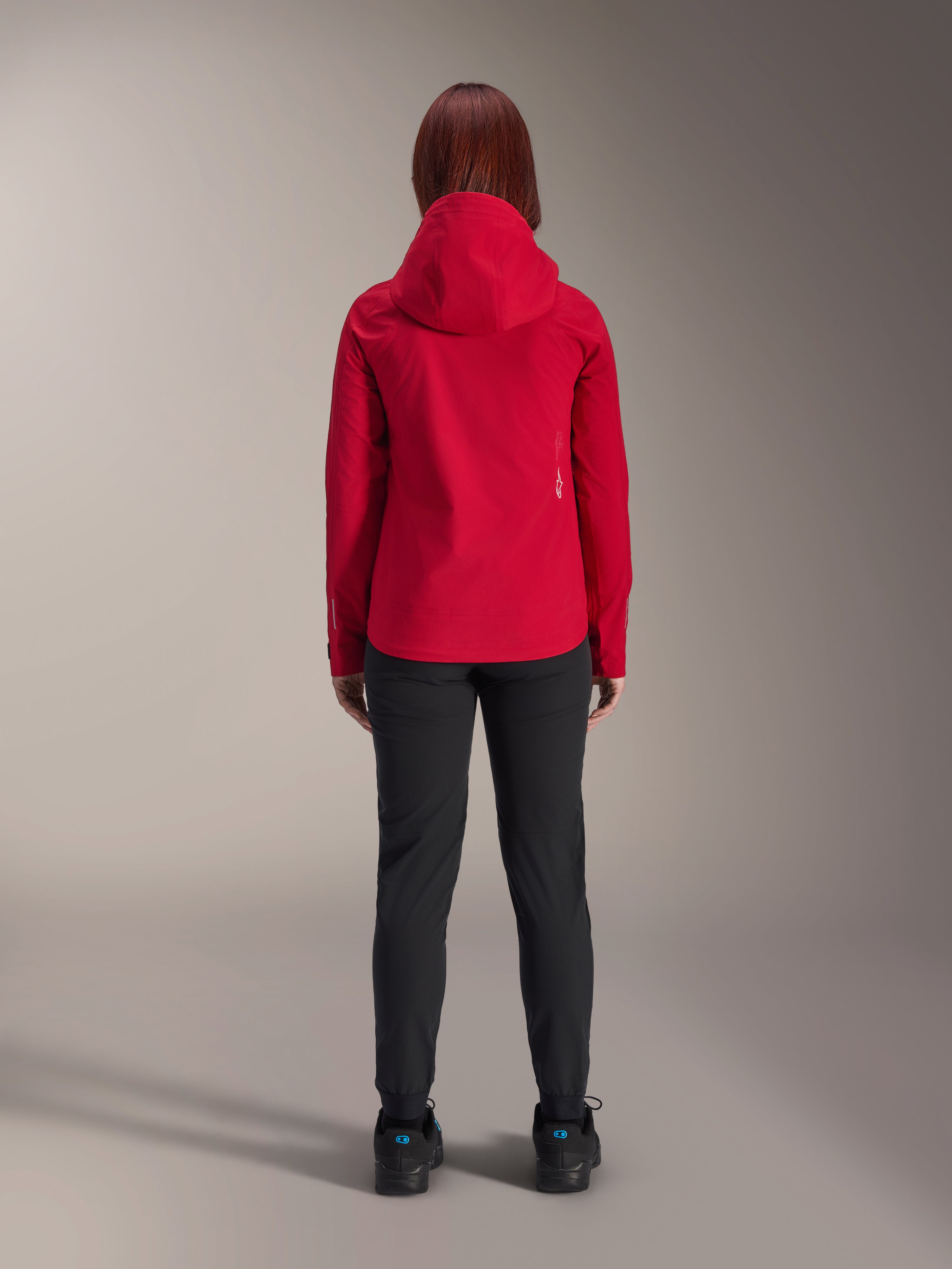 Achteraanzicht van een vrouw die de Alpinestars Stella A-Dura Elite Waterproof Jacket in Red Fluo draagt, gecombineerd met een zwarte broek en schoenen, voorzien van een beschermende capuchon en een verlengde achterzoom voor mountainbiken.