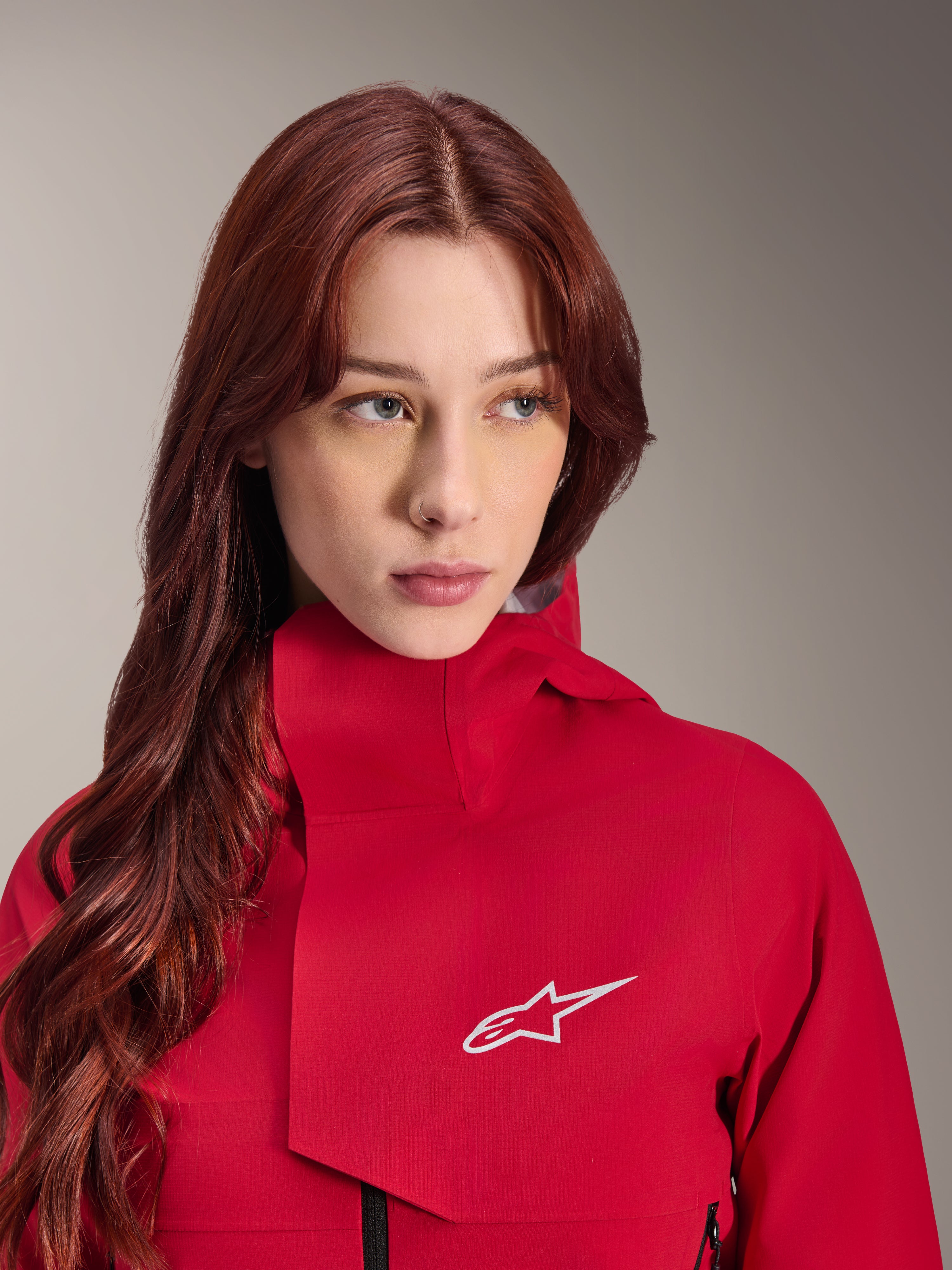 Alpinestars Stella A-Dura Elite Waterdichte Jas, Red Fluo, gedragen door een vrouw met lang rood haar, technische 3-laags waterdichte jas met hoge kraag en wit logo, hoogwaardige outdoor- en mountainbike-uitrusting