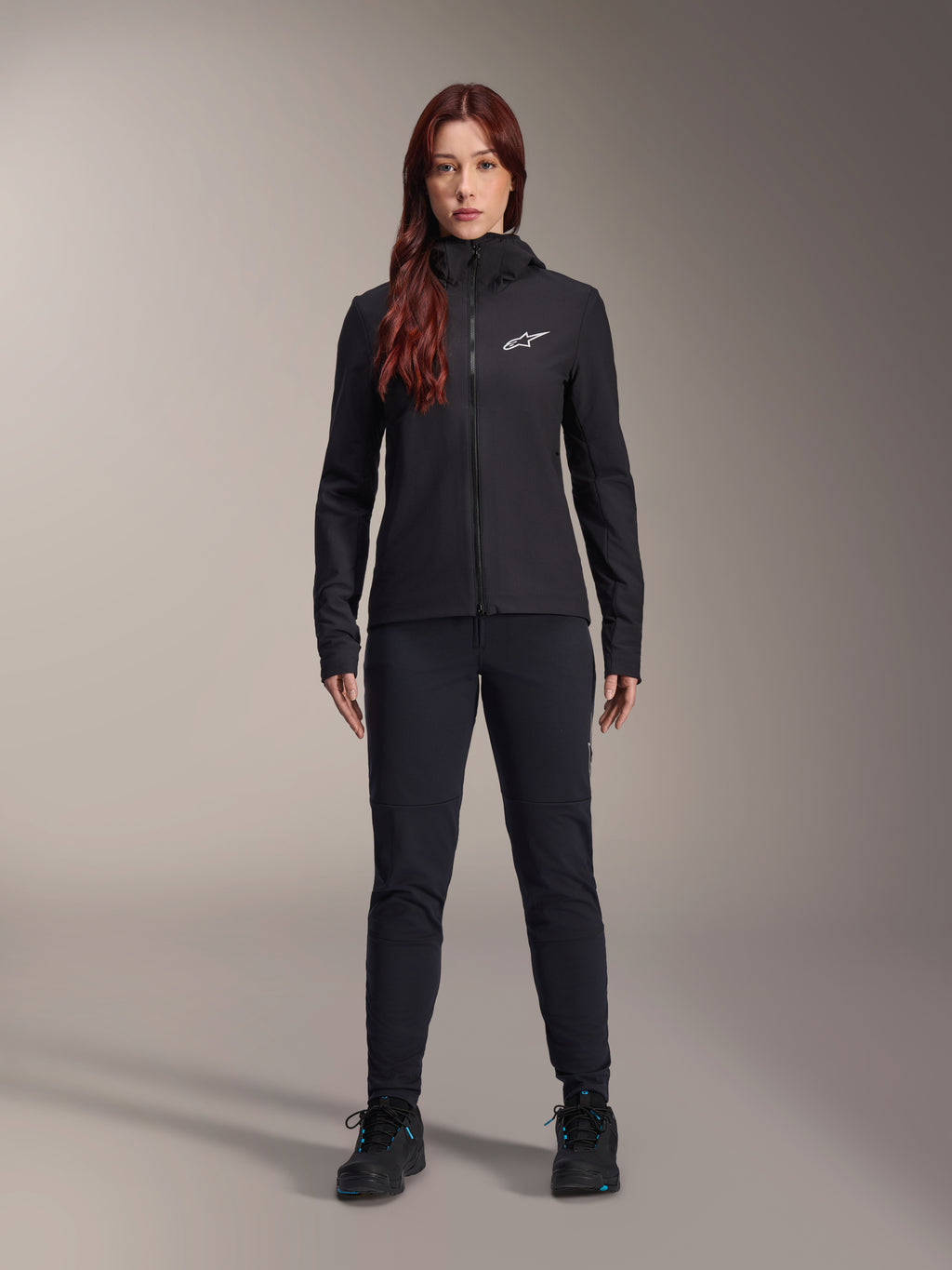 Alpinestars Stella A-Dura Thermal Jacket voor dames, zwarte lichtgewicht geïsoleerde fietsjas met capuchon, wit Astars-logo op de borst, gecombineerd met een zwarte broek en sportschoenen