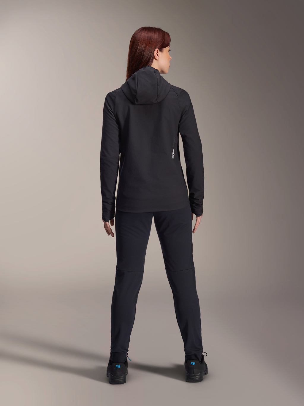 Vrouw draagt Alpinestars Stella A-Dura Thermal Jacket, achteraanzicht, zwarte geïsoleerde fietsjas met capuchon, bijpassende zwarte broek en schoenen, voorzien van een klein wit logo op de onderrug