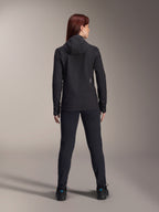 Vrouw draagt Alpinestars Stella A-Dura Thermal Jacket, achteraanzicht, zwarte geïsoleerde fietsjas met capuchon, bijpassende zwarte broek en schoenen, voorzien van een klein wit logo op de onderrug