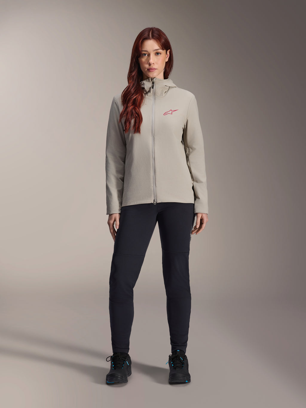 Alpinestars Stella A-Dura Thermal Jacket voor dames, mountainbikejas, titaniumgrijs, gedragen met een zwarte technische broek en zwarte schoenen, voorzien van een met fleece gevoerde capuchon en een rood logo op de borst, ontworpen voor ritten in koud weer