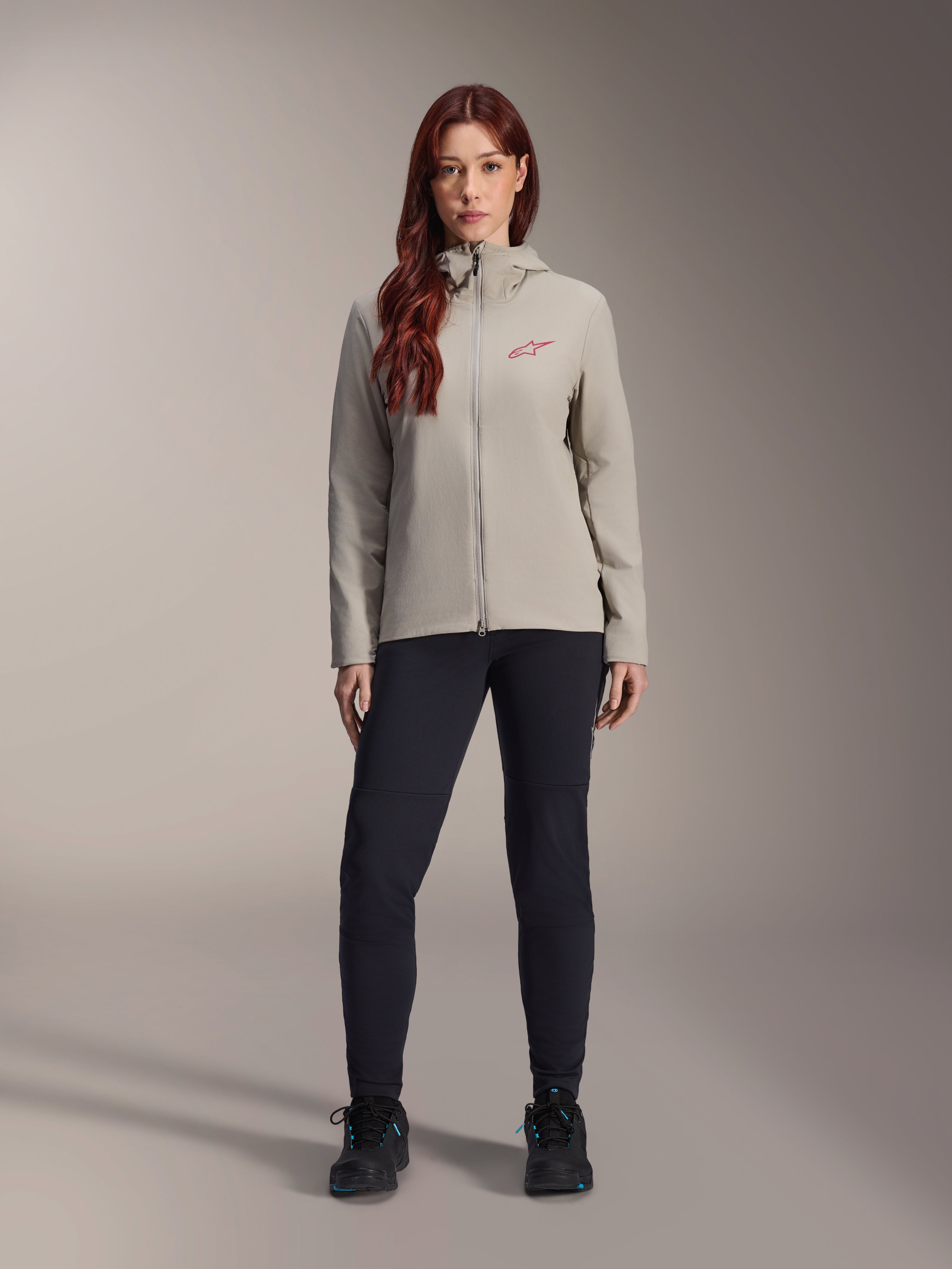 Alpinestars Stella A-Dura Thermal Jacket voor dames, mountainbikejas, titaniumgrijs, gedragen met een zwarte technische broek en zwarte schoenen, voorzien van een met fleece gevoerde capuchon en een rood logo op de borst, ontworpen voor ritten in koud weer