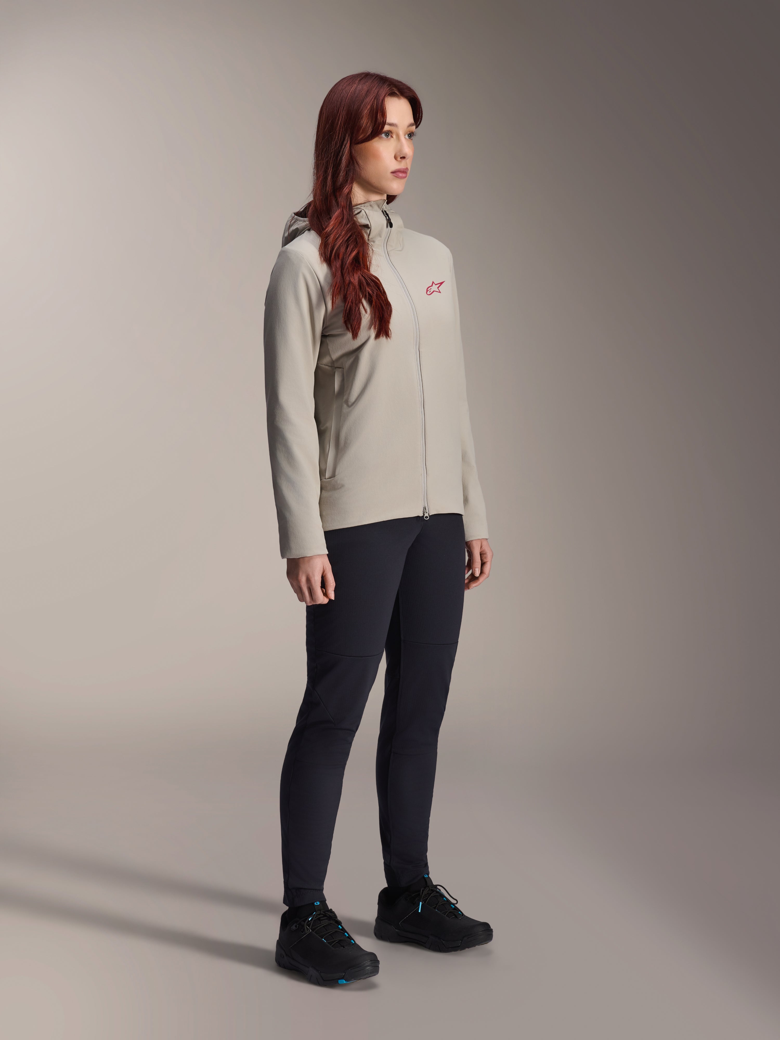 Alpinestars Stella A-Dura Thermal Jacket, titaniumgrijs, thermische fietsjas voor dames met een rood sterlogo op de borst, gecombineerd met een zwarte technische broek en zwarte mountainbikeschoenen, studio-opname voor outdoor-fietskleding