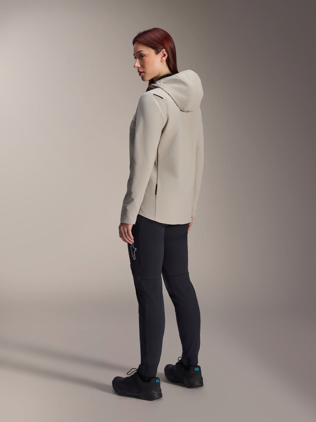 Alpinestars Stella A-Dura Thermal Jacket in Titanium grijs, driekwart achteraanzicht van een vrouw die de geïsoleerde fietsjas met capuchon draagt met een zwarte MTB-broek, studio-opname die de normale pasvorm en het zakdetail toont.