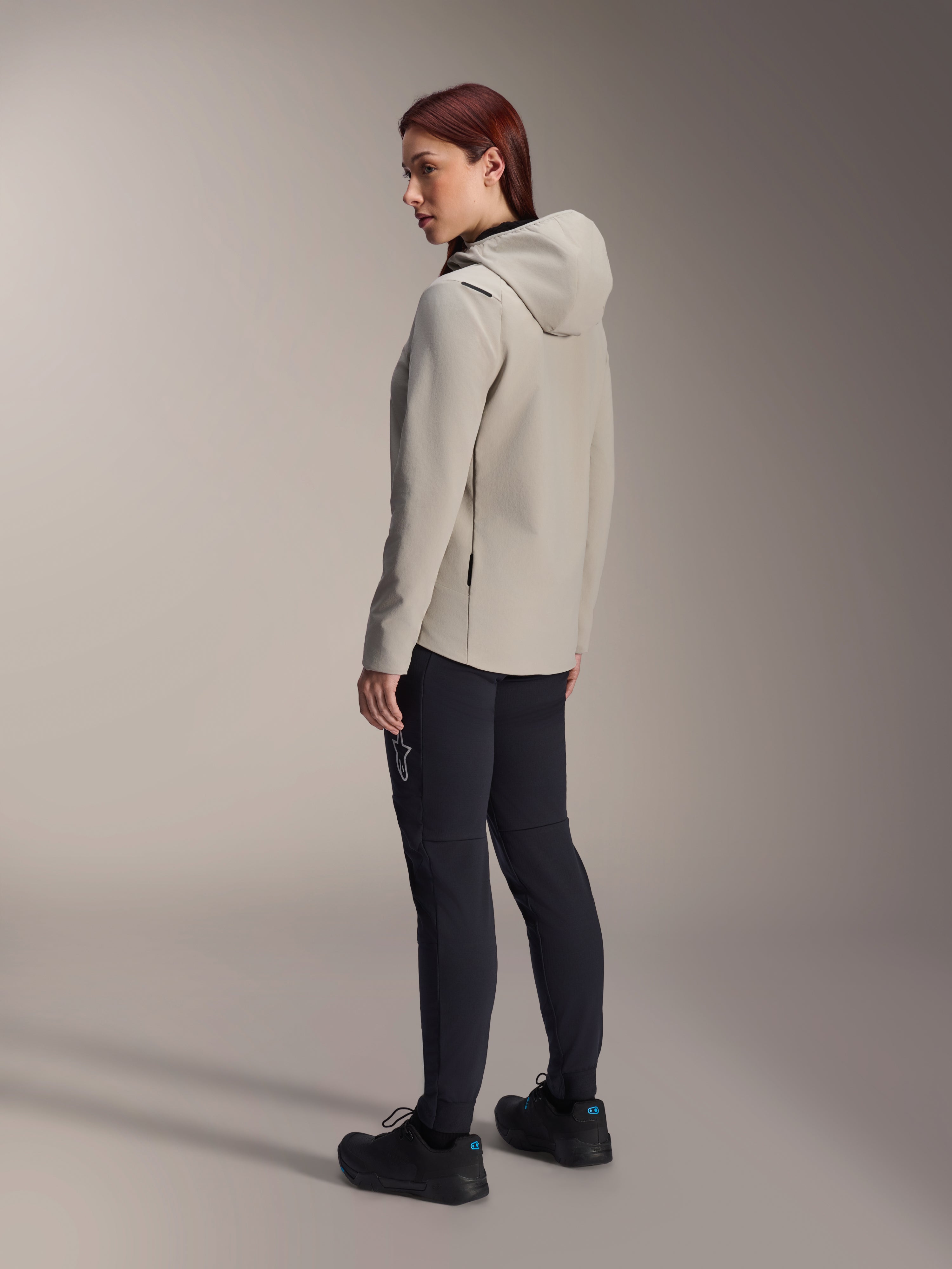 Alpinestars Stella A-Dura Thermal Jacket in Titanium grijs, driekwart achteraanzicht van een vrouw die de geïsoleerde fietsjas met capuchon draagt met een zwarte MTB-broek, studio-opname die de normale pasvorm en het zakdetail toont.