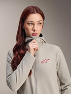 Alpinestars Stella A-Dura Thermojas voor dames, titaniumgrijs met een roze Alpinestars-sterlogo, model met lang rood haar die de jas met capuchon draagt en dichtritst, lichtgewicht geïsoleerde fietskleding voor koud weer.