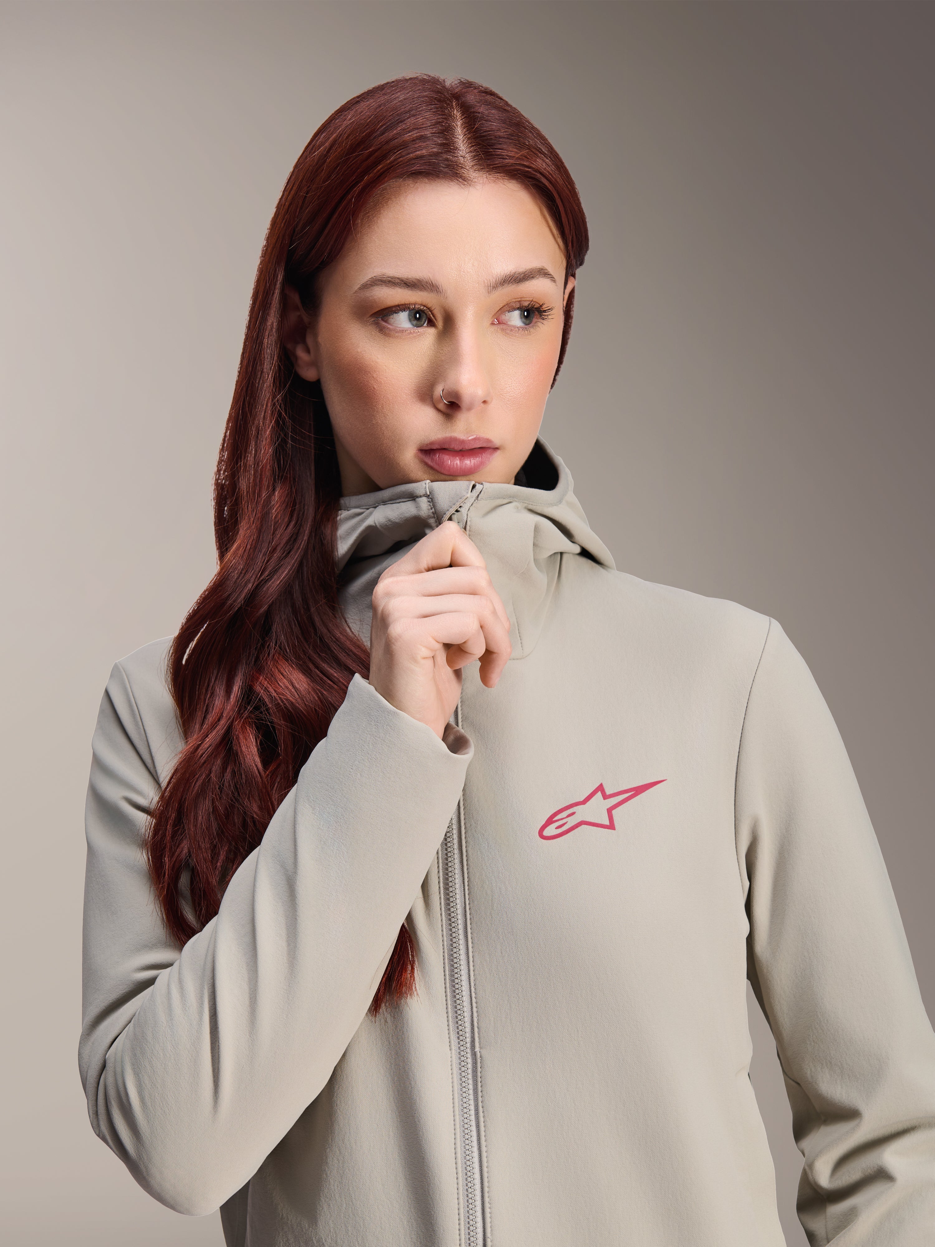 Alpinestars Stella A-Dura Thermojas voor dames, titaniumgrijs met een roze Alpinestars-sterlogo, model met lang rood haar die de jas met capuchon draagt en dichtritst, lichtgewicht geïsoleerde fietskleding voor koud weer.