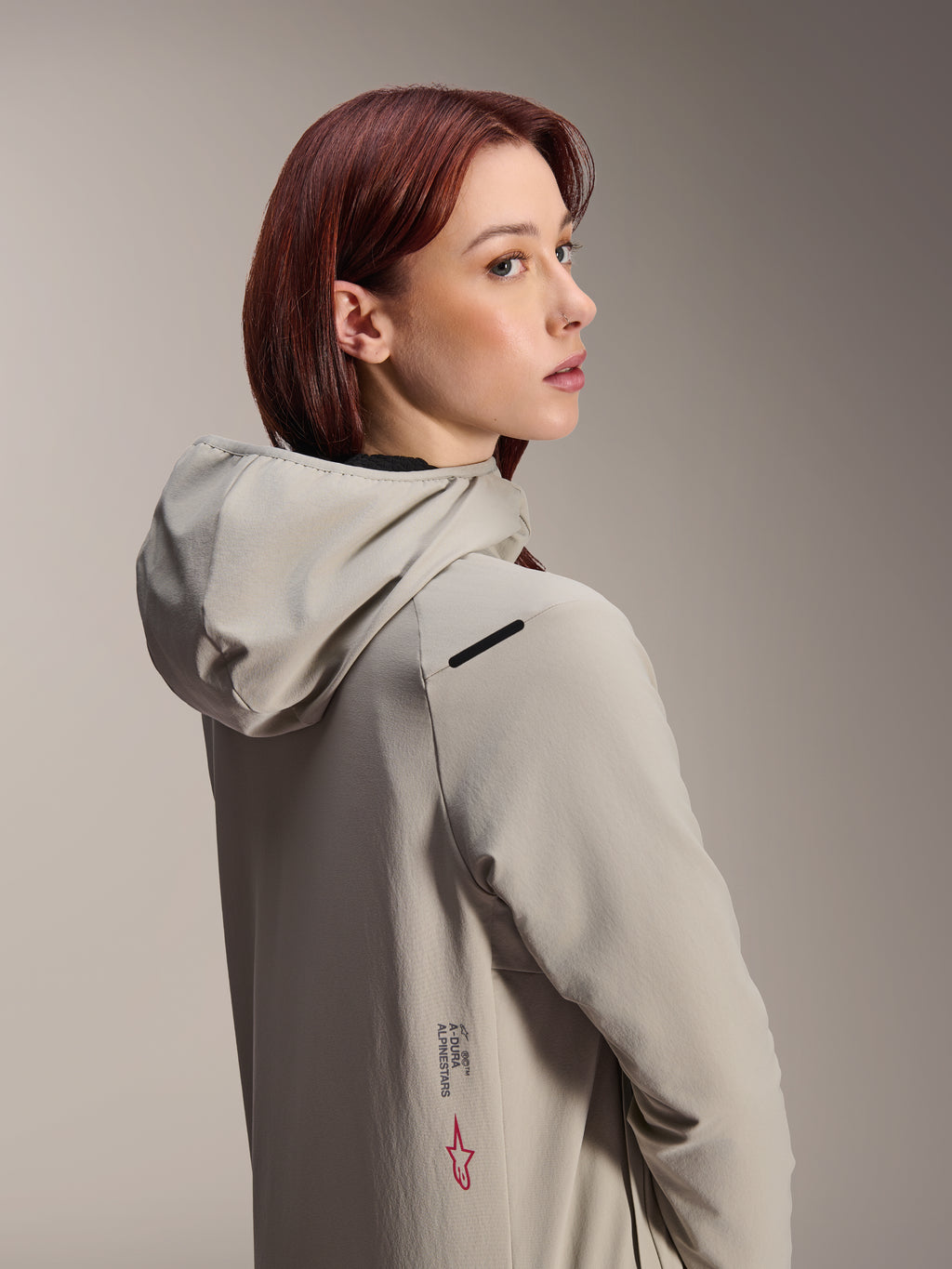Alpinestars Stella A-Dura Thermal Jacket, MTB-jas voor dames, Titanium grijs, vrouw die over haar schouder kijkt met een technisch jack met capuchon, voorzien van Alpinestars-tekst en rood sterlogo op de zijkant, ontworpen voor fietsen in koud weer