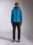 Model wearing the Alpinestars Speeder Chromium Windbreak Jacket in Blue, featuring a hooded design with black drawcords, a black front zipper, and a black Astars chest logo, paired with black pants and sneakers, casual motorsport streetwear