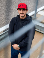 Man die de Alpinestars Overhaul Racer Jacket in het zwart draagt, een waterbestendig regenjack met capuchon en een klein logo op de borst, gecombineerd met een rode Alpinestars pet, casual motorsport lifestyle kleding