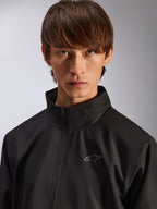 Alpinestars Overhaul Racer Jacket, zwarte regenjas, close-up portret van een mannelijk model dat de jas draagt, met een hoge kraag en een klein zilveren Astars-logo op de borst, motorsport-geïnspireerde casual kleding