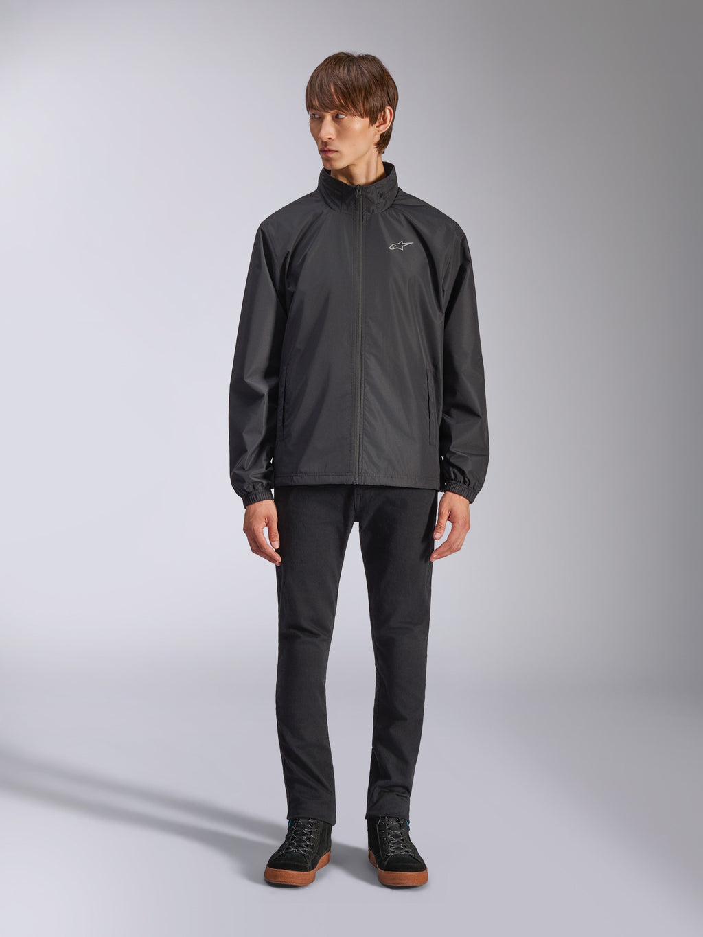 Alpinestars Overhaul Racer Jacket, zwarte regenjas voor heren, volledige weergave op een model gecombineerd met een zwarte broek en casual sneakers, voorzien van een klein wit logo op de borst, waterbestendige stof en een casual op motorsport geïnspireerd ontwerp