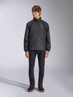 Alpinestars Overhaul Racer Jacket, zwarte regenjas voor heren, volledige weergave op een model gecombineerd met een zwarte broek en casual sneakers, voorzien van een klein wit logo op de borst, waterbestendige stof en een casual op motorsport geïnspireerd ontwerp