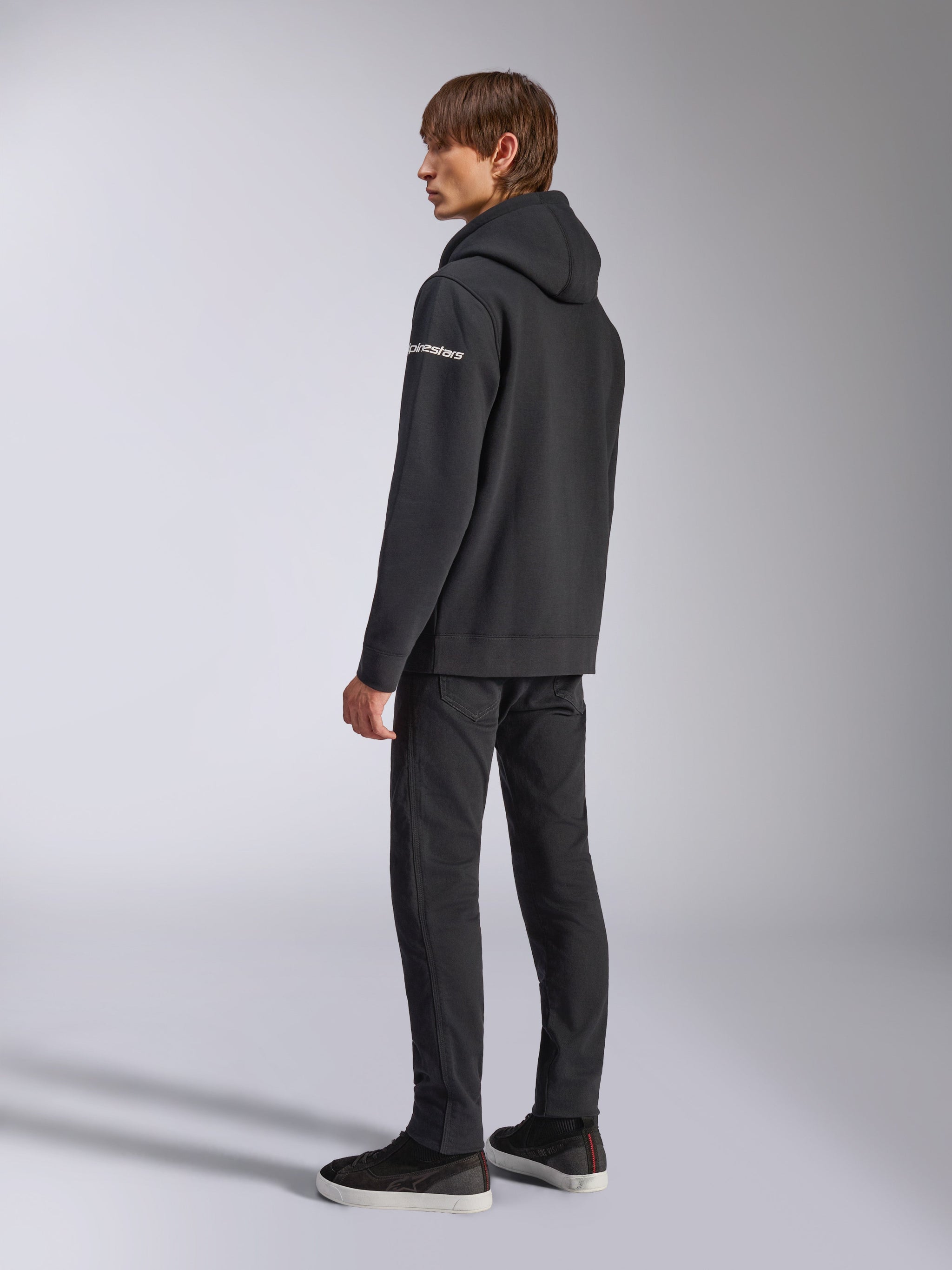 Alpinestars Frontal Hoodie, casual streetwear, zwart, achteraanzicht van opzij met wit woordmerklogo op de mouw, gedragen met zwarte broek en sneakers, modern motorsport-geïnspireerd ontwerp