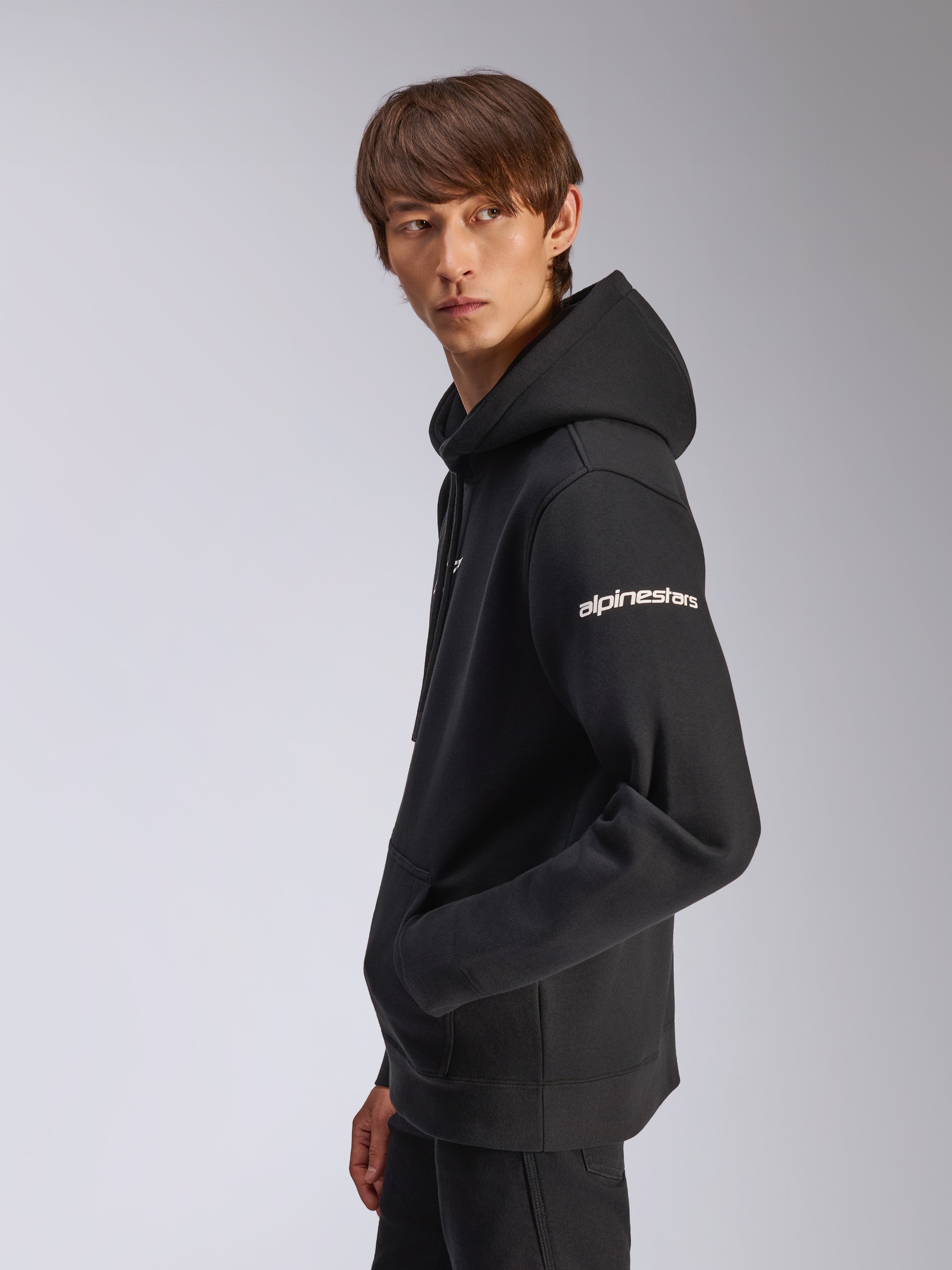 Alpinestars Frontal Hoodie, zwarte pullover fleece, model draagt item in zijaanzicht, wit Alpinestars woordmerk op de mouw, kangoeroezak, casual motorsport-geïnspireerde streetwear