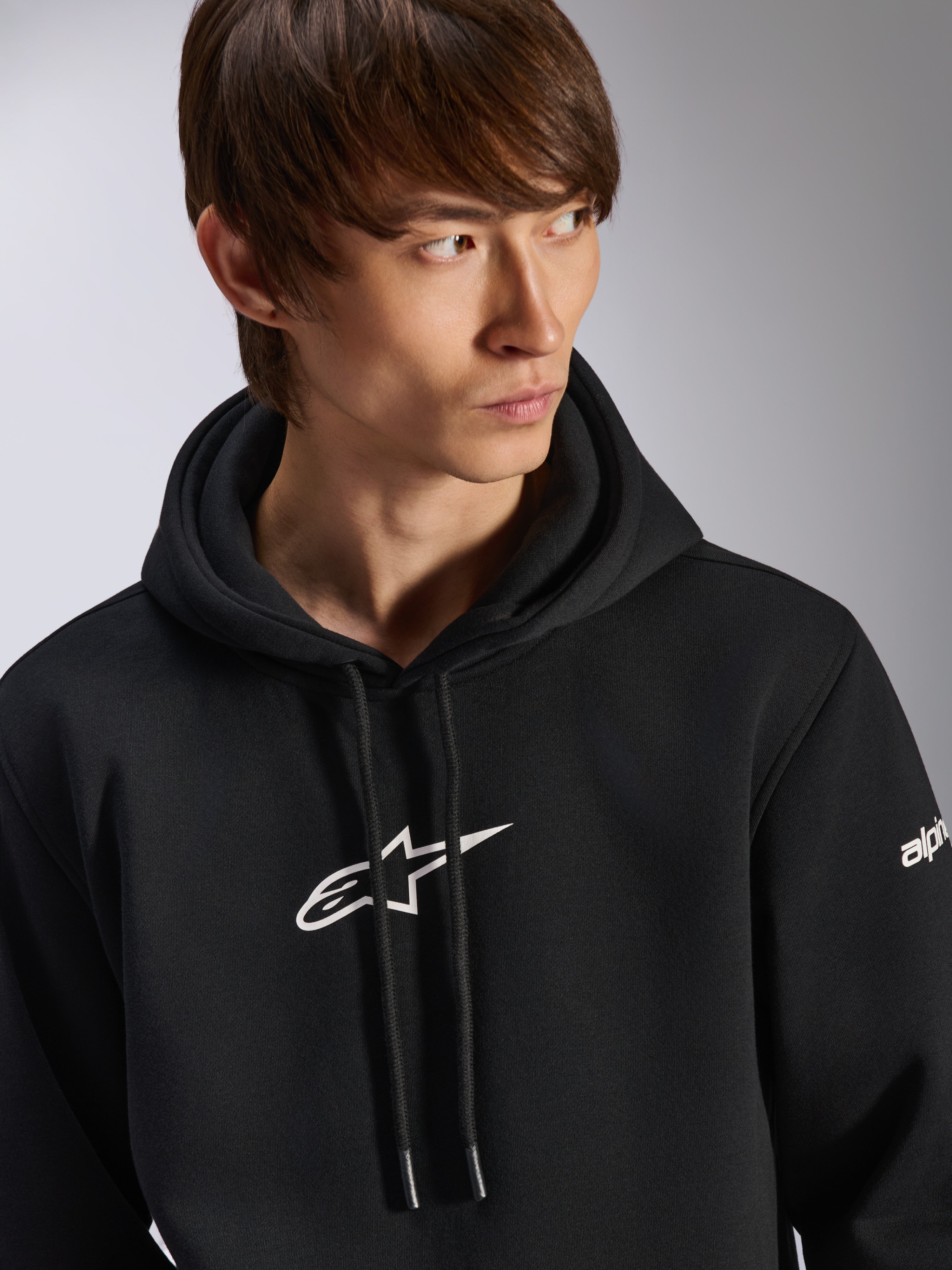 Alpinestars Frontal Hoodie, zwarte pullover fleece hoodie, wit Astar-logo op de borst en woordmerk op de mouw, capuchon met trekkoord, gedragen door mannelijk model, casual motorsport streetwear