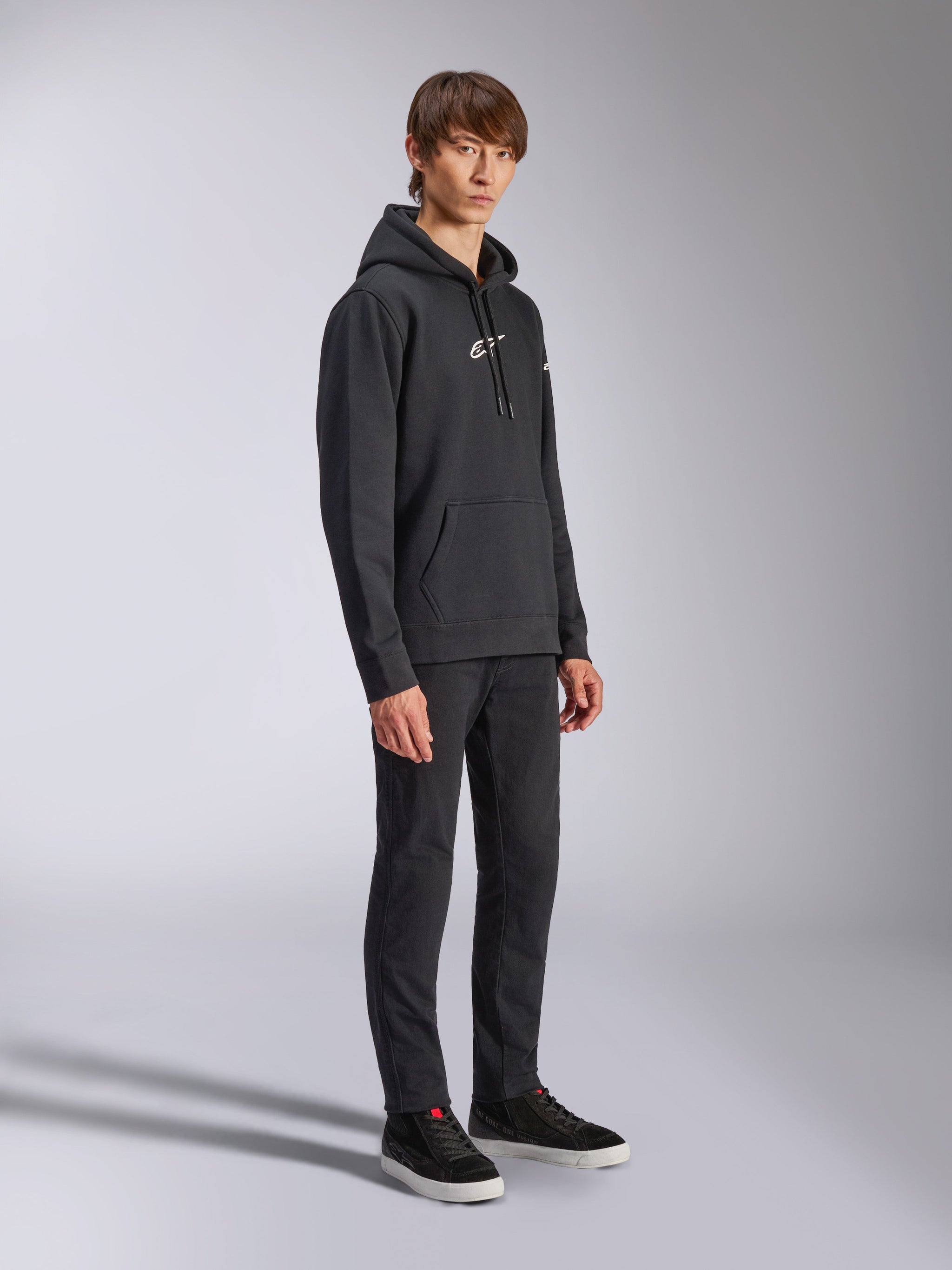 Alpinestars Frontal Hoodie, zwart, pullover fleece hoodie met wit HD-logo op de borst en woordmerklogo's op de mouwen, kangoeroezak, gecombineerd met een zwarte broek en sneakers, casual motorsport-geïnspireerde streetwear