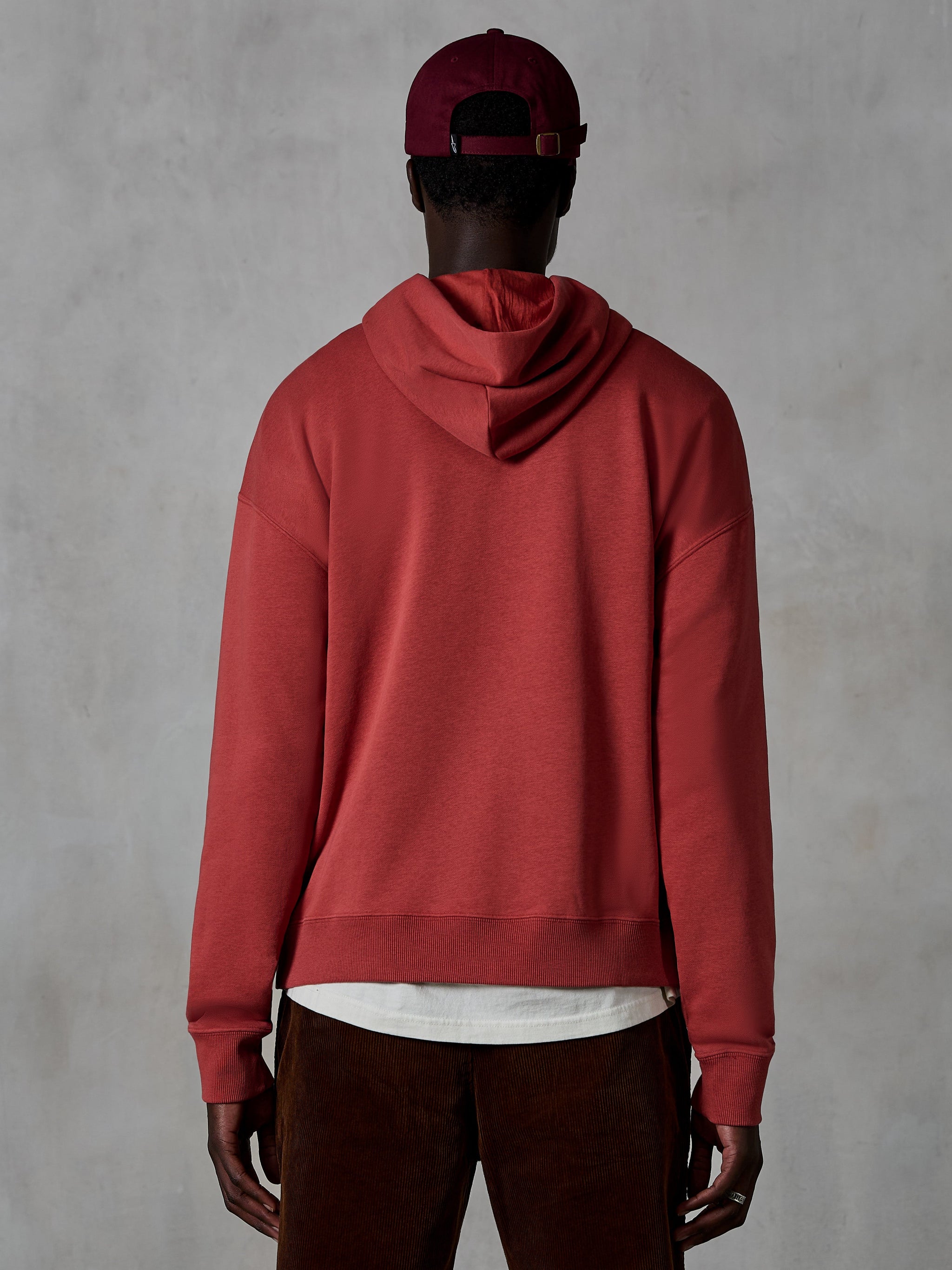Alpinestars Essentials Ageless Hoodie, casual fleece hoodie, Brick rood, achteraanzicht van model in lifestyle kleding, voorzien van een strak ontwerp met geribbelde zoom en manchetten, gecombineerd met een bruine corduroy broek en een bordeauxrode pet