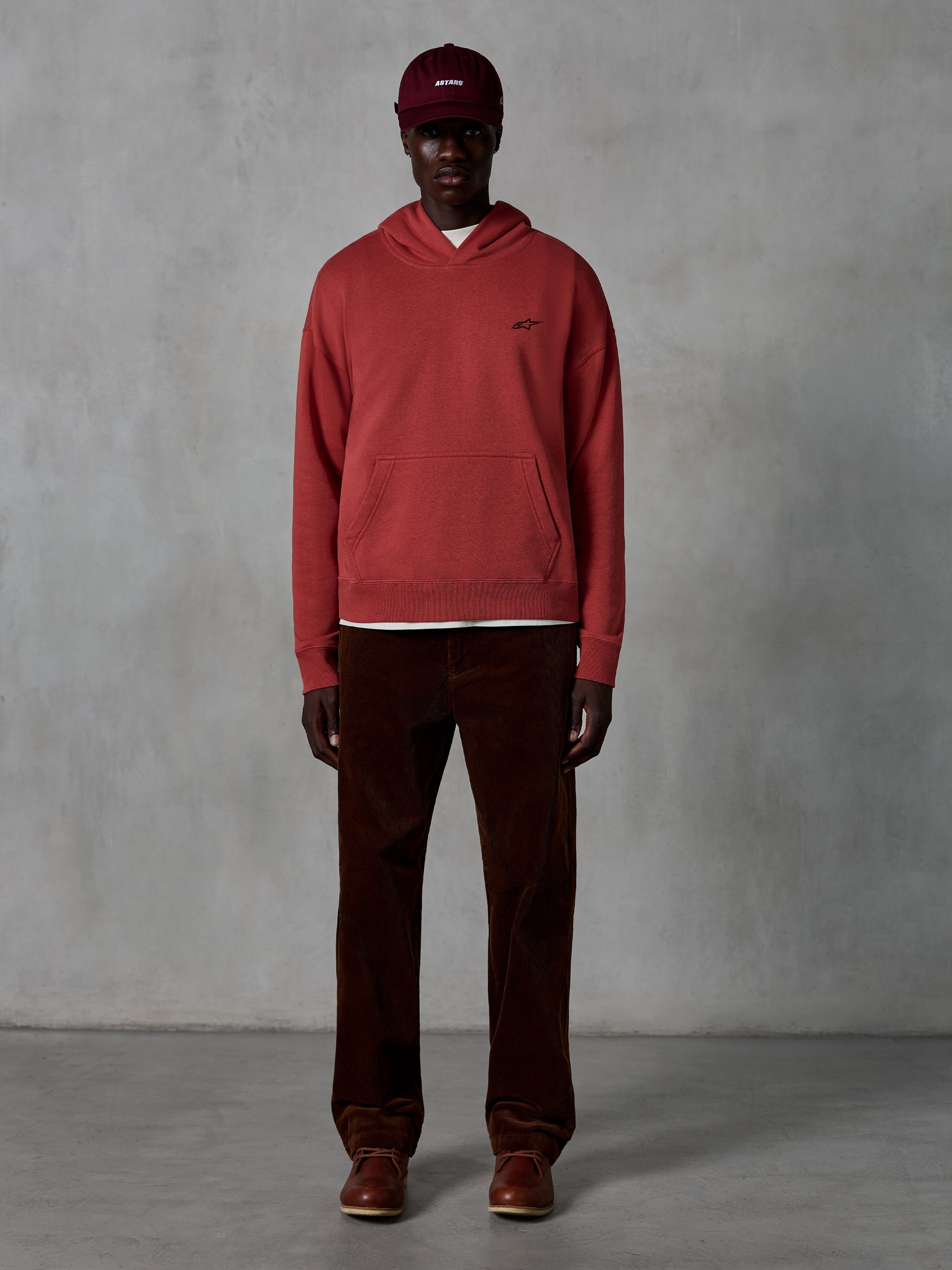 Alpinestars Essentials Ageless Hoodie, casual sportswear hoodie in Brick rood met een garment stone wash-afwerking, voorzien van een klein zwart Alpinestars-logo op de borst en een kangoeroezak aan de voorzijde, gedragen door een model met een bordeauxrode pet en bruine corduroy broek voor een strakke streetwear-look.