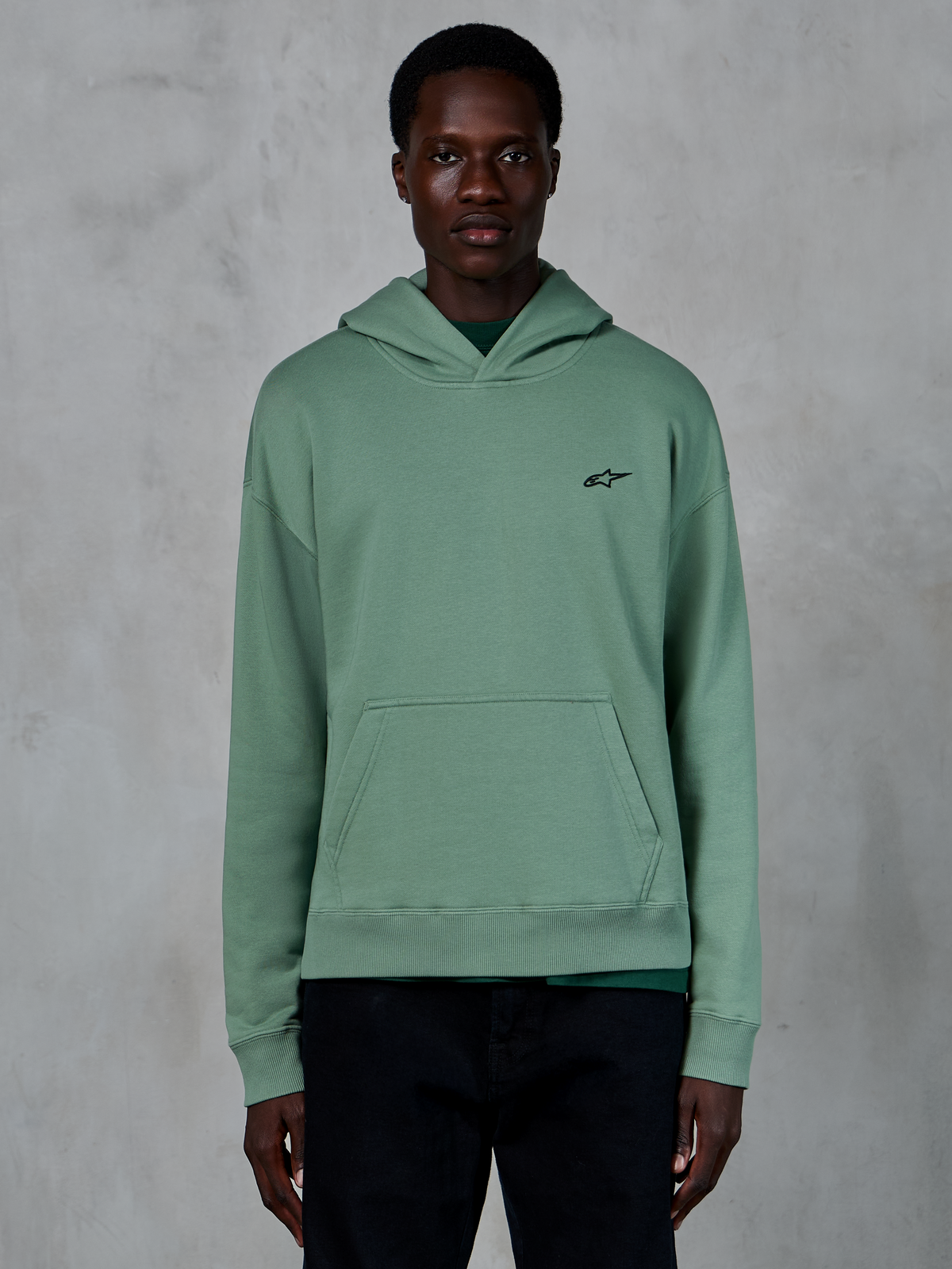 Alpinestars Essentials Ageless Hoodie, cactusgroen, casual fleece hoodie met een stone-washed finish, voorzien van een klein zwart sterlogo op de borst en een kangoeroezak aan de voorkant, gedragen door een model in een strakke lifestyle-setting