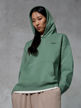 Alpinestars Essentials Ageless Hoodie, casual streetwear, cactusgroen met klein zwart logo, hoodie gedragen met de capuchon omhoog, kangoeroezak aan de voorkant, stone-washed afwerking, gecombineerd met een lichtbeige broek