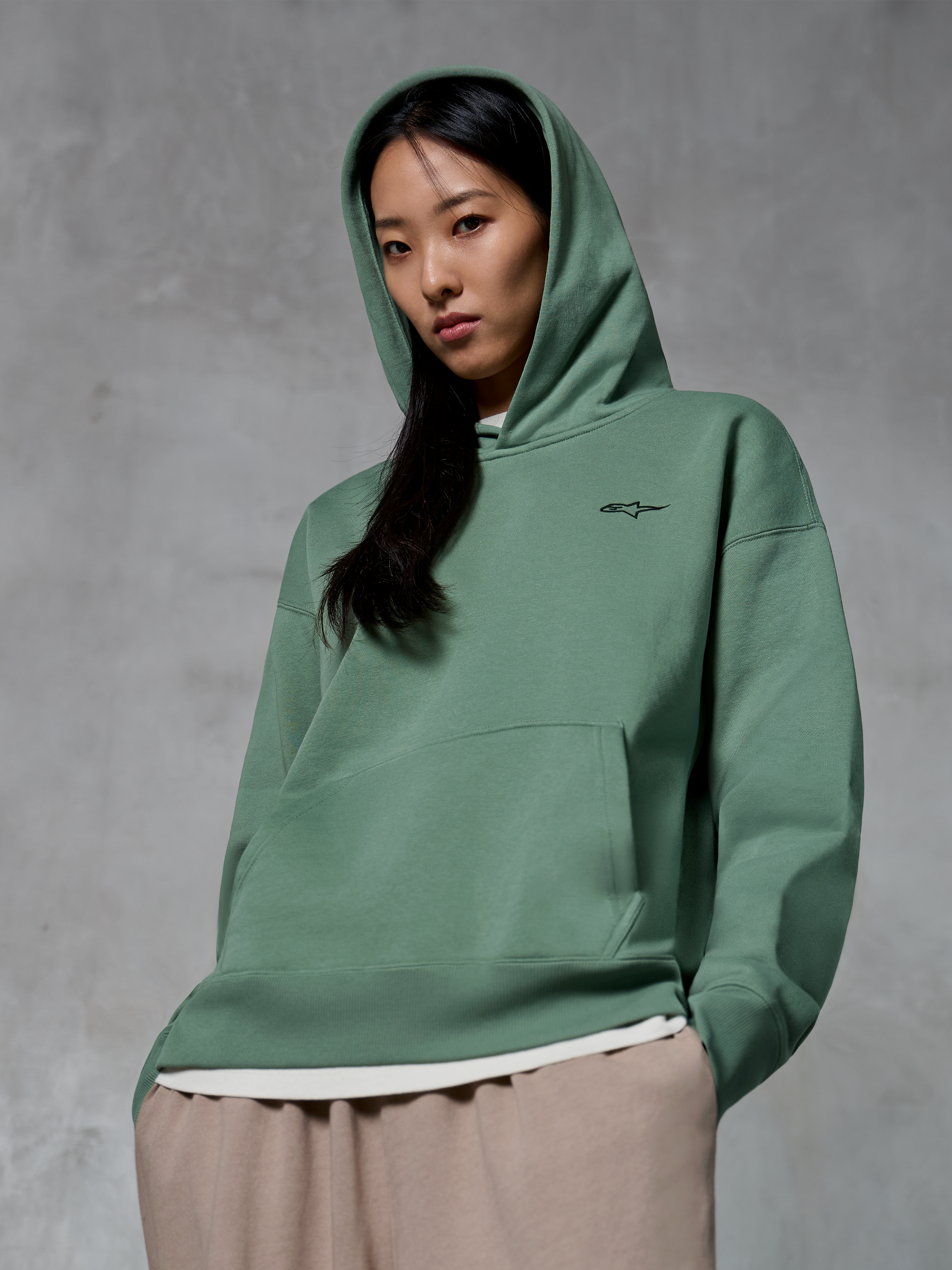 Alpinestars Essentials Ageless Hoodie, casual streetwear, cactusgroen met klein zwart logo, hoodie gedragen met de capuchon omhoog, kangoeroezak aan de voorkant, stone-washed afwerking, gecombineerd met een lichtbeige broek