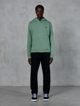 Alpinestars Essentials Ageless Hoodie, cactusgroen, klein zwart logo op de borst, kangoeroezak, casual lifestyle streetwear gedragen met een zwarte broek en zwart-witte sneakers tegen een muur met textuur